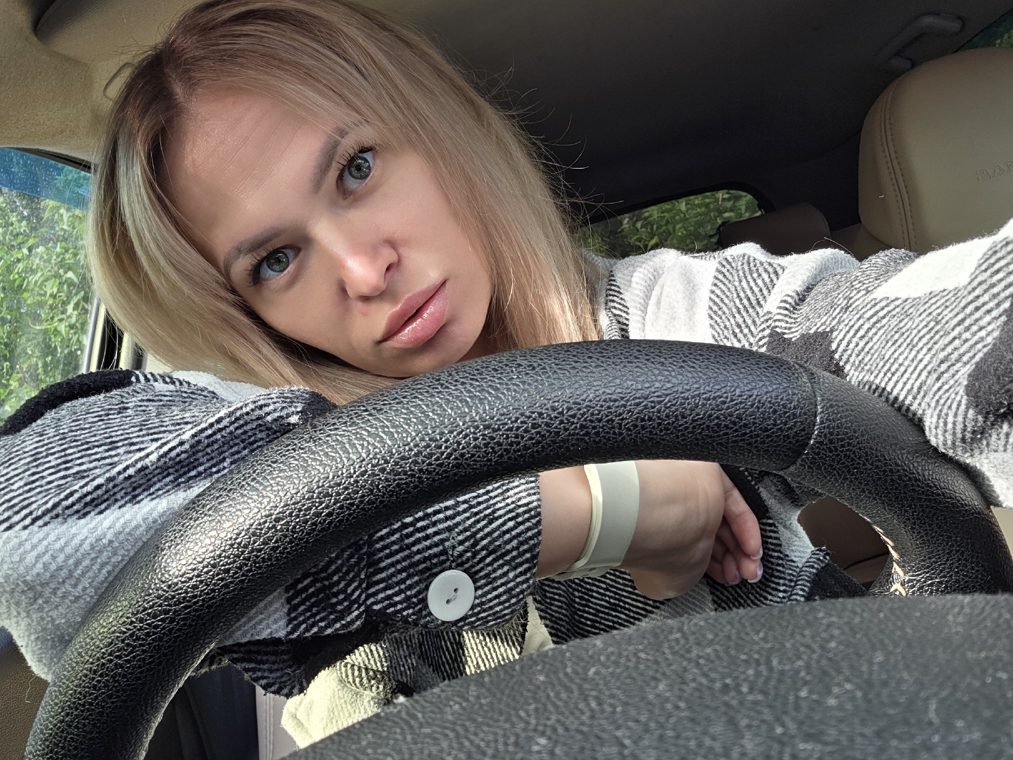 A woman with blonde hair and blue eyes is in the driver's seat of a car. She is wearing a black and white plaid shirt. Her left arm is resting on the steering wheel, and she is looking at the camera. The interior of the car is visible, including the dashboard, steering wheel, and a headrest. Green foliage can be seen through the car window. The image appears to have been taken in Chaikovsky, Russia.
