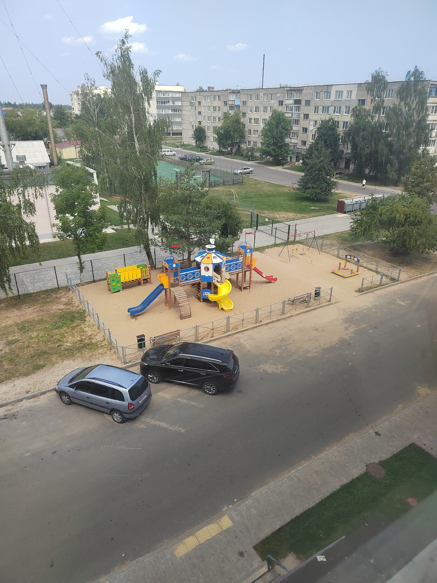 An aerial view captures a residential area with apartment buildings, a playground, and parked cars on a paved road. The scene is set in the city of Slawharad, Belarus. In the foreground, two cars are parked on the asphalt road; a silver station wagon on the left and a black SUV on the right. Adjacent to the road is a fenced playground filled with sand, featuring a colorful play structure with slides, swings, and climbing elements. Beyond the playground, a green grassy area is visible. In the background, several multi-story apartment buildings dominate the skyline, typical of Soviet-era architecture, interspersed with trees. The sky is partly cloudy, indicating daytime. No people are clearly visible in the immediate vicinity of the playground or cars. The overall impression is that of a typical urban residential environment on a calm day.
