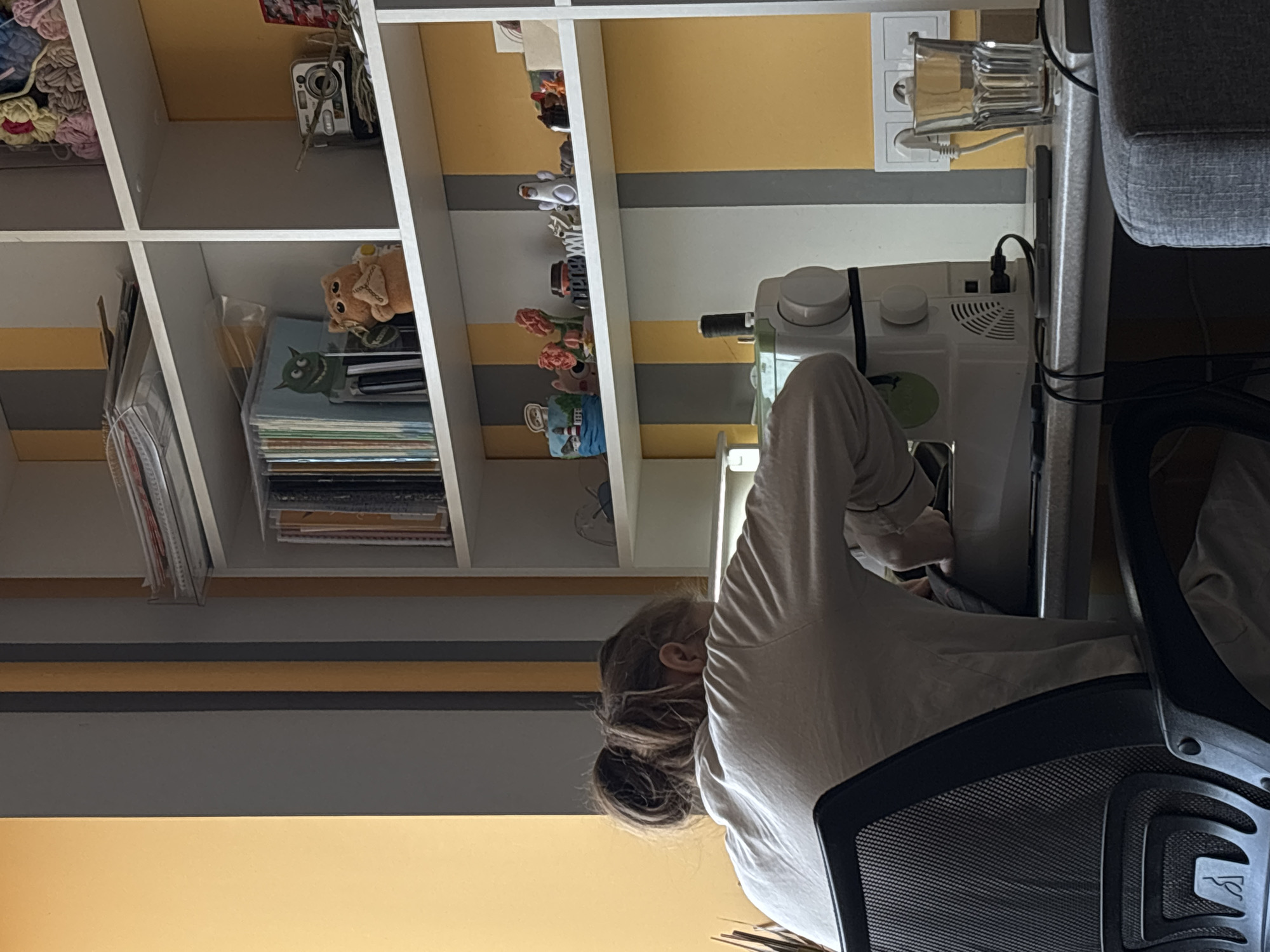 A woman is seated in a black mesh office chair, operating a white sewing machine. The sewing machine is positioned on a table against a wall with horizontal gray and yellow stripes. Behind her is a white bookshelf, containing items such as crocheted yarn, a vintage camera, small figurines, and stacks of paper or books. An electrical outlet and a water glass are visible above the sewing machine. The location is likely in Perm, Russia, given the image context.