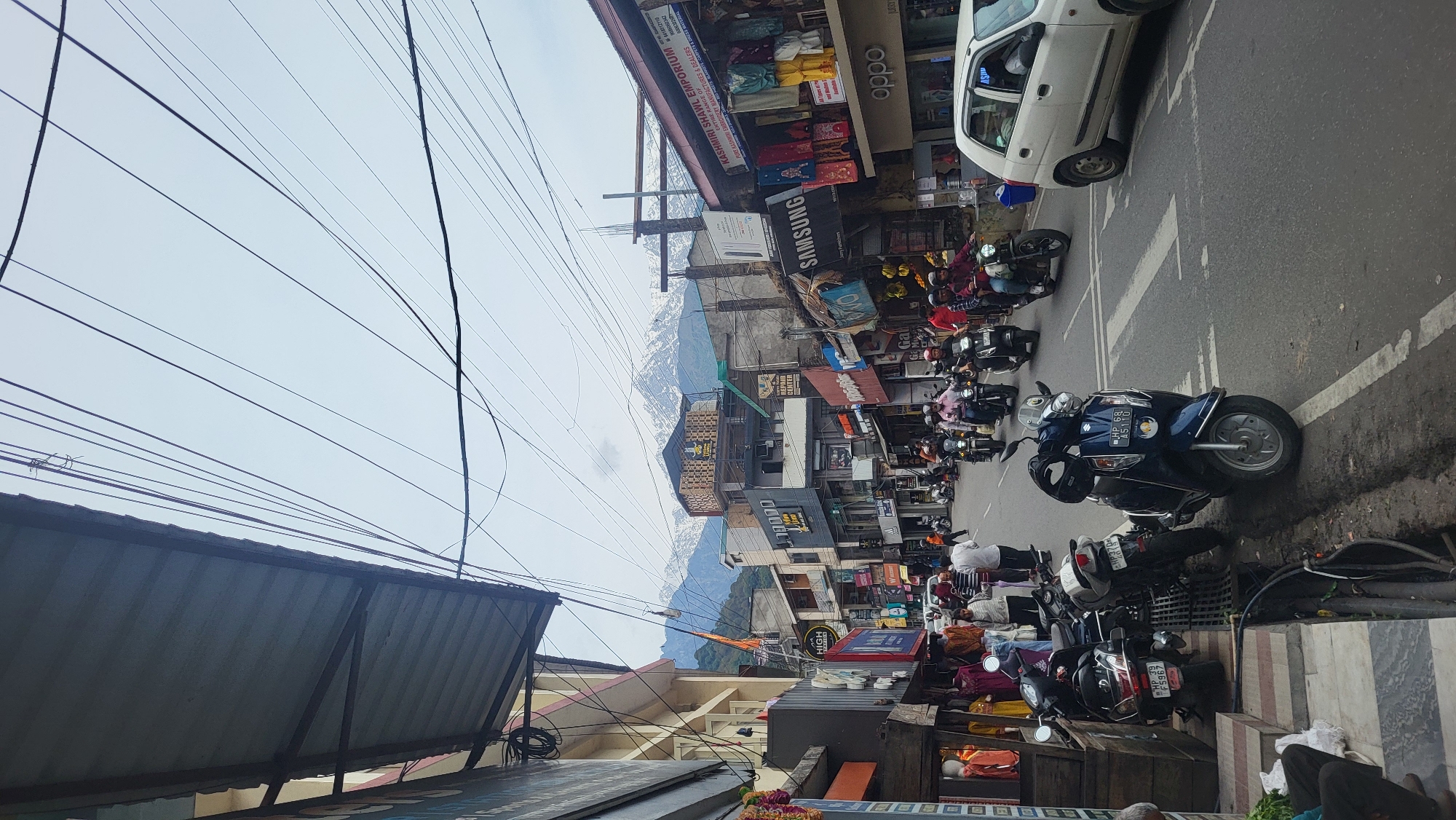 The image captures a bustling street scene in Dharamshala, India. Numerous scooters and motorbikes are parked along the roadside and traveling along the right side of the road. A white car is parked near a building. Shops line the street on both sides, with signs visible for brands such as "Samsung" and "Oppo," as well as local businesses like "Kashmiri Shawl Emporium." In the background, mountains are visible through gaps between buildings. Overhead, a dense network of power lines crisscrosses the sky. People appear to be moving along the sidewalks and interacting with the businesses.