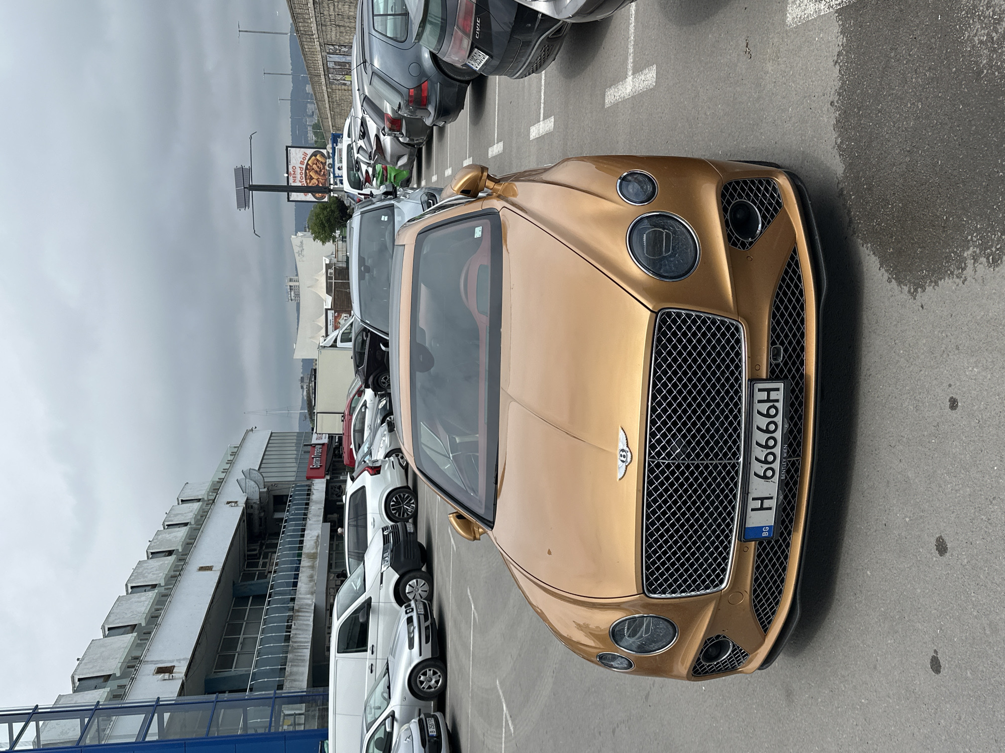 An overhead shot captures a striking gold Bentley parked in an outdoor asphalt parking lot under an overcast sky. The luxury car, likely a Continental GT, is angled towards the viewer, prominently displaying its distinctive grille, circular headlights, and the iconic "B" emblem. Its Bulgarian license plate reads "BG H 66666H", with "BENTLEY WIEN" visible underneath, suggesting its origin.
Behind the Bentley, a diverse row of other parked vehicles, including various sedans, SUVs, and white vans, occupies marked spaces. White parking lines segment the asphalt, which exhibits several damp patches, suggesting recent rain. No people are visible in the scene.
In the background, a large, modern building with a multi-level facade and extensive glass sections features a red sign that clearly says "Бургас Моторс" (Burgas Motors). Further away, a billboard advertises "NEMO Seafood Boil". Distant hills or mountains complete the horizon under the cloudy conditions, indicating a coastal or hilly region.
The image depicts a daytime scene with a grey, cloudy sky, suggesting a cool or mild weather day. The Bulgarian license plates and the "Бургас Моторс" sign confirm the location in Bulgaria, consistent with the provided context of Varna. The overall scene is quiet, with parked cars as the primary subjects in an urban or commercial setting.