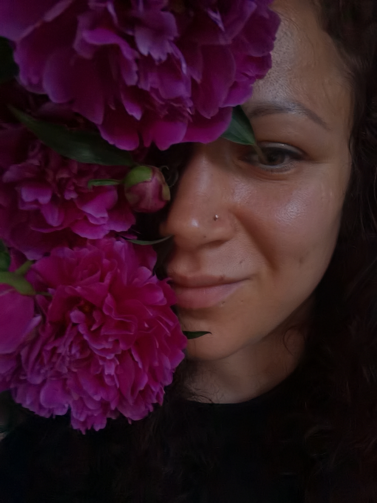 A medium shot captured from a slightly low angle reveals a woman's face partially obscured by large, vibrant pink peonies. She has curly dark hair, dark eyes, and a nose piercing. Her skin appears warm-toned and smooth, with subtle highlights on her cheeks and forehead. One eye is visible, looking towards the left of the frame, while her mouth is slightly parted in a soft smile. Her right cheek displays a dimple. The background is dark, with the peonies occupying the left and upper portions of the image, their petals showing intricate textures and shades of pink and magenta. The composition creates a sense of intimacy and focus on the woman and the flowers. There is no information or visual cue within the image to indicate a location in Chaikovsky, Russia.