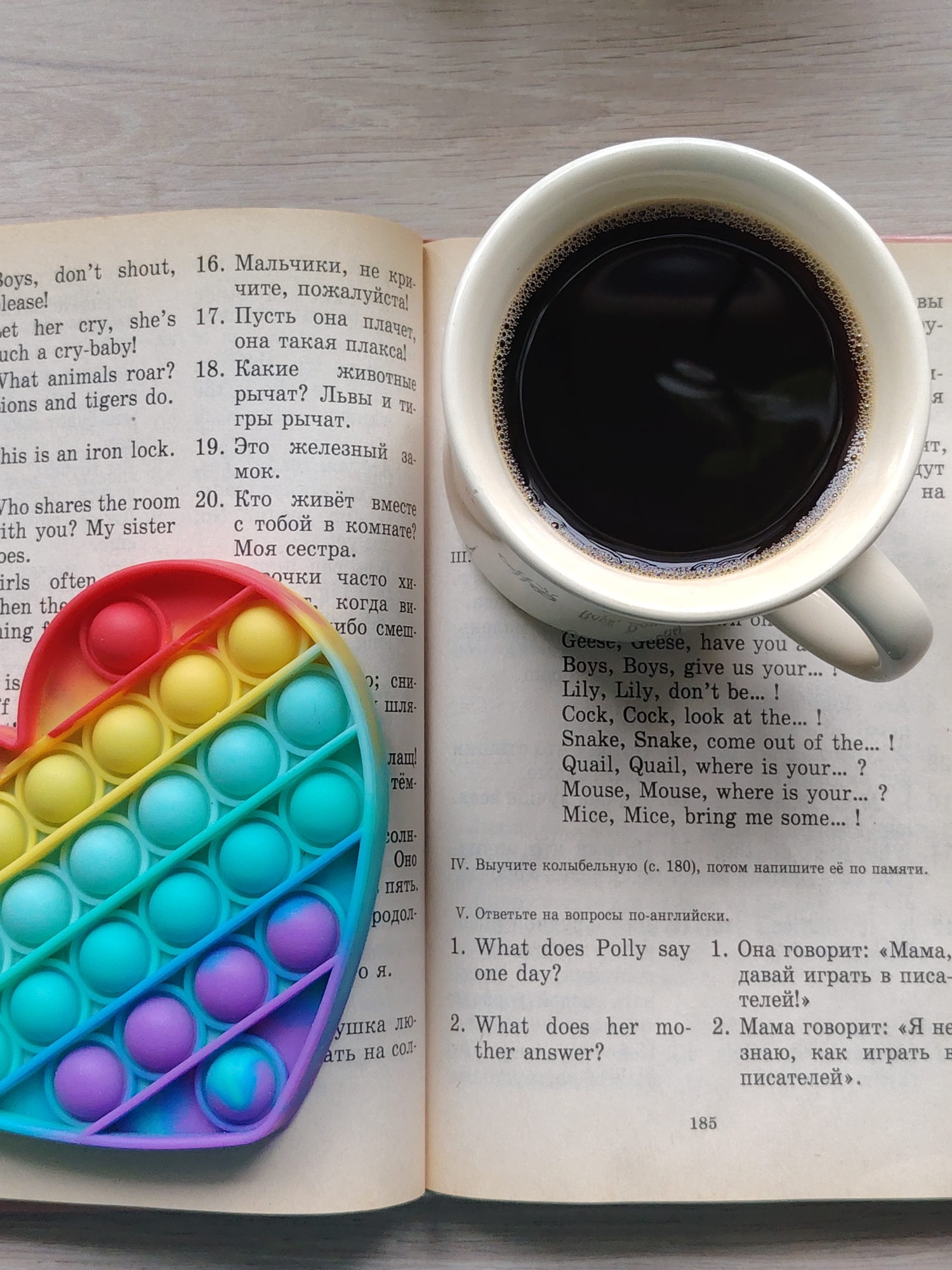 The image shows an open book with Russian and English text, a heart-shaped rainbow-colored fidget toy, and a mug of dark coffee. The book appears to be an English learning textbook. The scene conveys a sense of studying and relaxation. Location information (Chaikovsky, Russia) is not discernible within the image content.