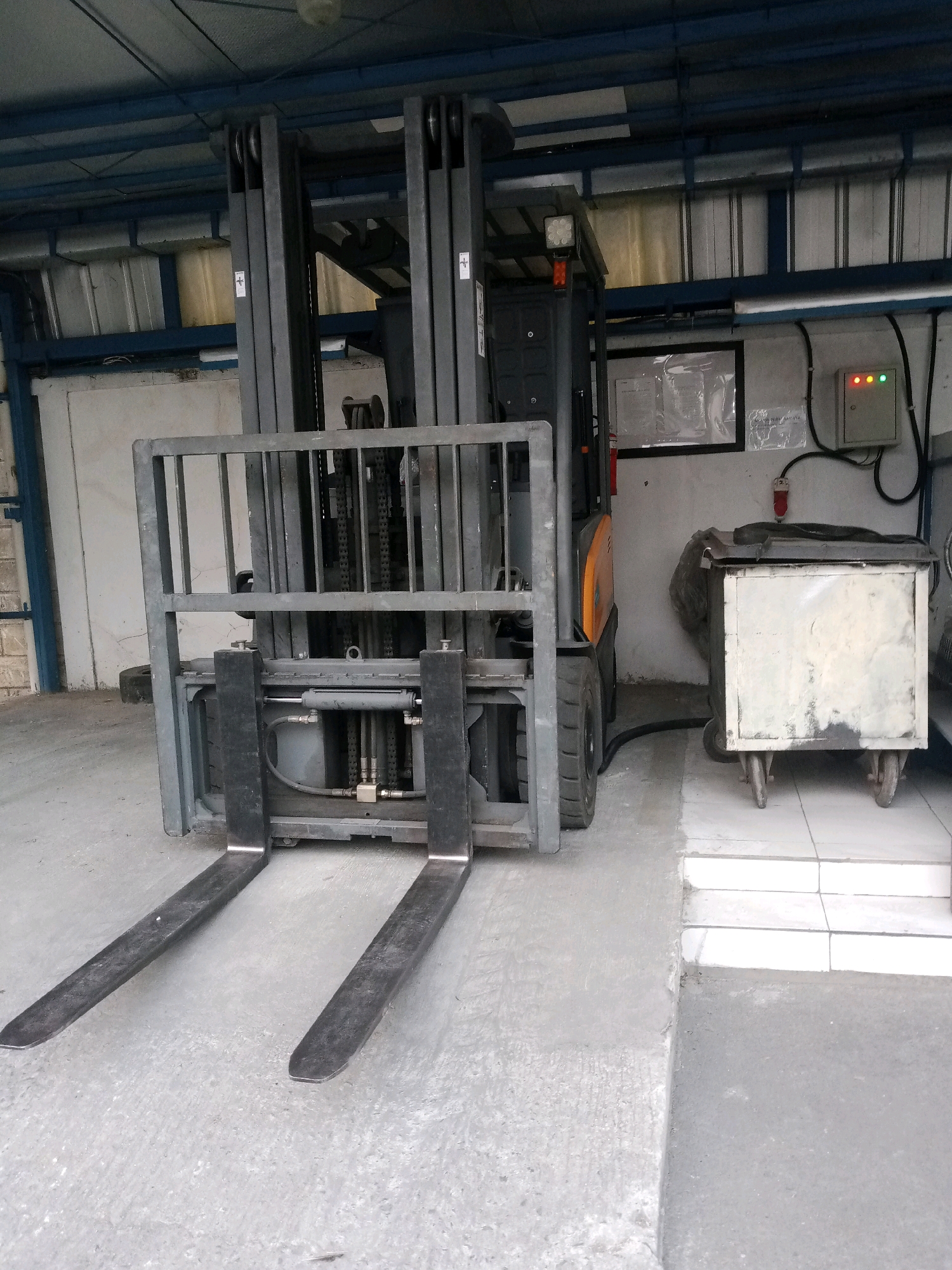 This media file depicts a forklift parked inside a warehouse or industrial building. The forklift is the primary subject, a dark gray industrial vehicle with prominent yellow accents on its body. Its forks are extended forward, resting on a concrete floor. To the right of the forklift, there is a white wheeled bin with a dark lid, also parked on the floor. In the background, metal shelving units and industrial lighting are visible. A control panel with three illuminated lights (red, yellow, and green) is mounted on the wall, along with some framed documents. The scene suggests an operational or recently used industrial environment. The lighting appears to be artificial, typical of an indoor space. The location context provided is Balaraja, Indonesia, which could imply the typical architecture and construction styles of such buildings in that region. No people are visible in the frame. There is no indication of the time of day or weather conditions. The overall mood is functional and industrial. Some faint text is visible on a document in the background, but it is illegible.