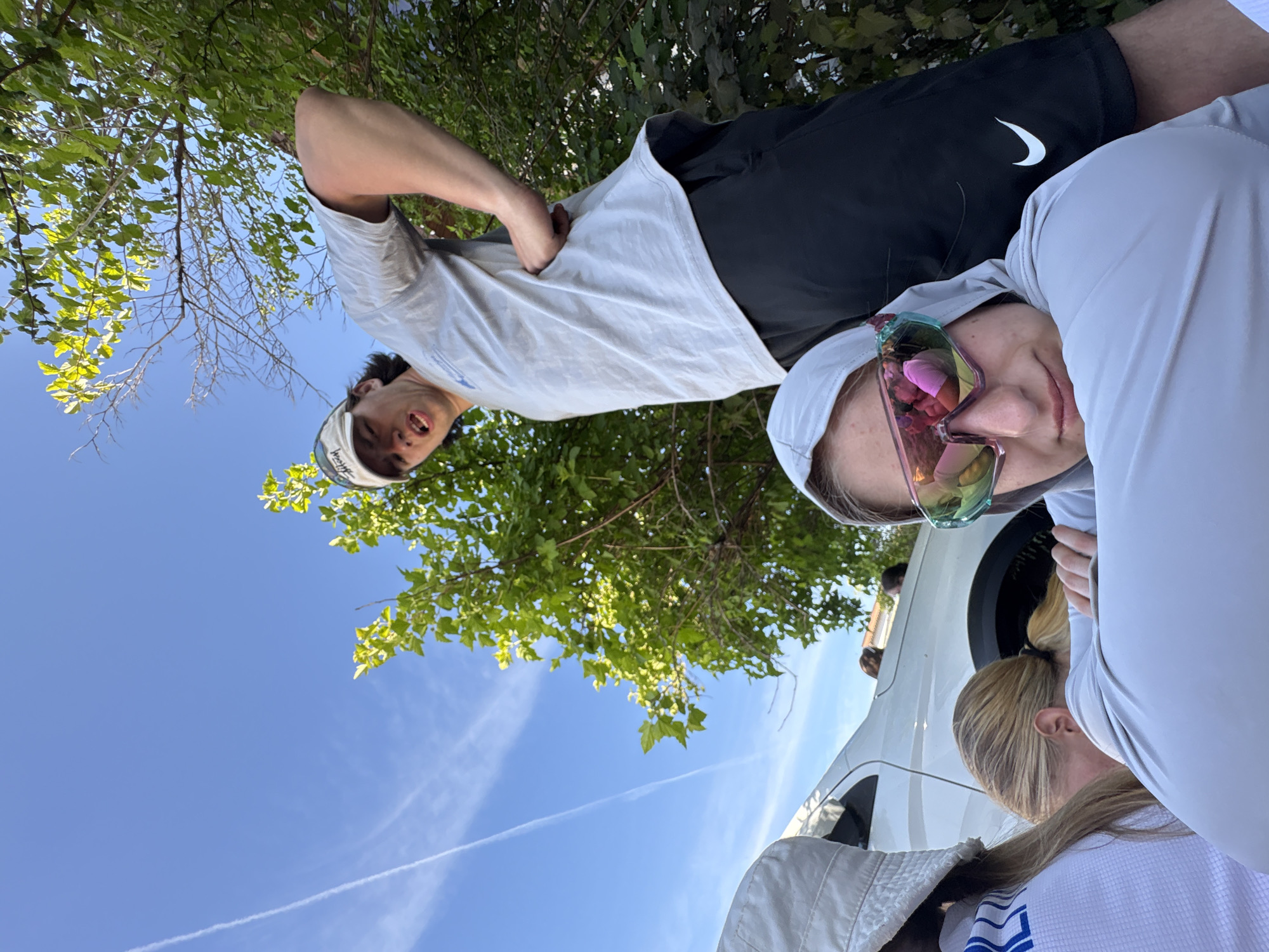 Two individuals are visible in the image. One appears to be a young man wearing a light-colored t-shirt, black shorts with a Nike logo, and a baseball cap. He has his arm in his pocket. The other individual is wearing a light-colored hooded top and iridescent sunglasses, and has blonde hair tied back in a ponytail. They appear to be standing near a white vehicle. The background includes green foliage and a blue sky with contrails. The location of Khvalynsk, Russia is not evidenced in the image.