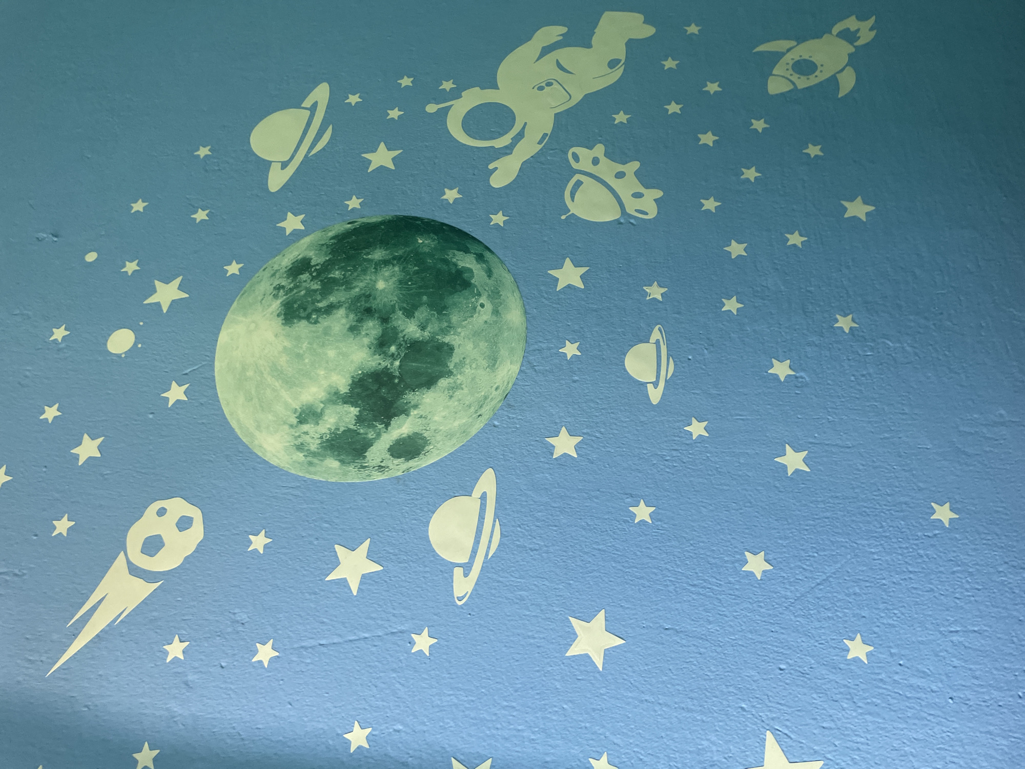 This is a close-up, eye-level shot of a blue wall decorated with glow-in-the-dark space-themed decals. The main subject is a large, detailed decal of the moon, prominently positioned in the left half of the frame. Surrounding the moon and scattered across the wall are numerous white decals depicting celestial bodies and space-faring objects. These include stars of various sizes, planets with rings, a rocket ship with flames, a comet with a tail, and an astronaut in mid-flight. There's also a decal of what appears to be a stylized acorn or a small, spherical planet with a crown-like ring. The decals are arranged in a whimsical, playful manner, suggesting a child's room or a themed play area. The blue of the wall has a slightly textured appearance, and the lighting in the image indicates it could be daytime, with the glow-in-the-dark feature not being actively illuminated. The scene is static, with no activity or event taking place, other than the implied movement of the celestial objects depicted by the decals. There is no visible text. The location context of Kołobrzeg, Poland, provides no direct visual cues within the image itself.