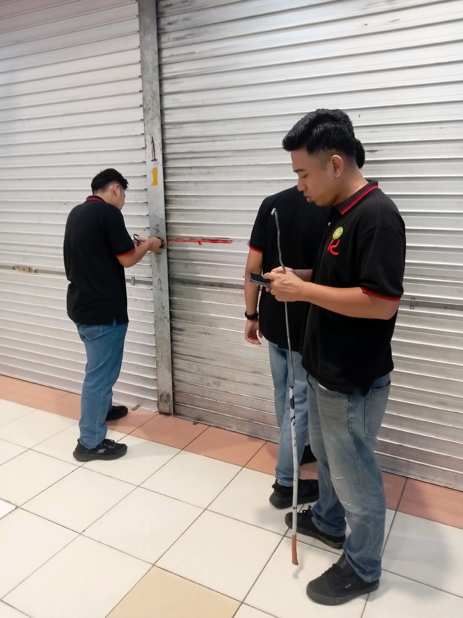 Three males are standing in front of a closed metallic roller door. The person in the foreground on the right is wearing a black polo shirt with red trim, blue jeans, and black sneakers. He is holding a smartphone in his left hand and a white cane with a brown tip in his right hand, which is resting on the floor. To his left, another male is wearing a black polo shirt and blue jeans, and he is holding a smartphone. Further to the left, a third male is wearing a black polo shirt and blue jeans. He is standing with his back to the camera and is interacting with the roller door, appearing to be measuring or marking it with a red tape measure. The roller door is closed and shows signs of wear. The floor is tiled with white and beige square tiles. The environment appears to be an indoor commercial space, possibly a shop or storefront, with the roller door suggesting it is a business. The presence of a cane may indicate that one of the individuals is visually impaired. The location is identified as Batam, Indonesia.