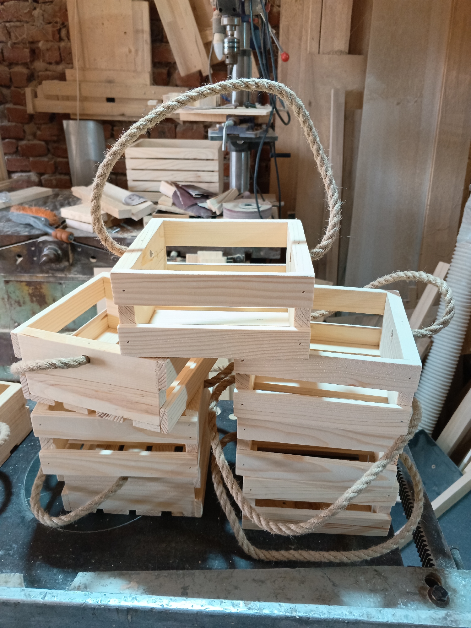 The image shows a stack of small, unfinished wooden crates with rope handles, likely handmade. The crates are arranged on what appears to be a workbench in a workshop setting. There are tools and woodworking equipment visible in the background, along with exposed brickwork on a wall. The city of Chaikovsky, Russia, cannot be inferred from the image.