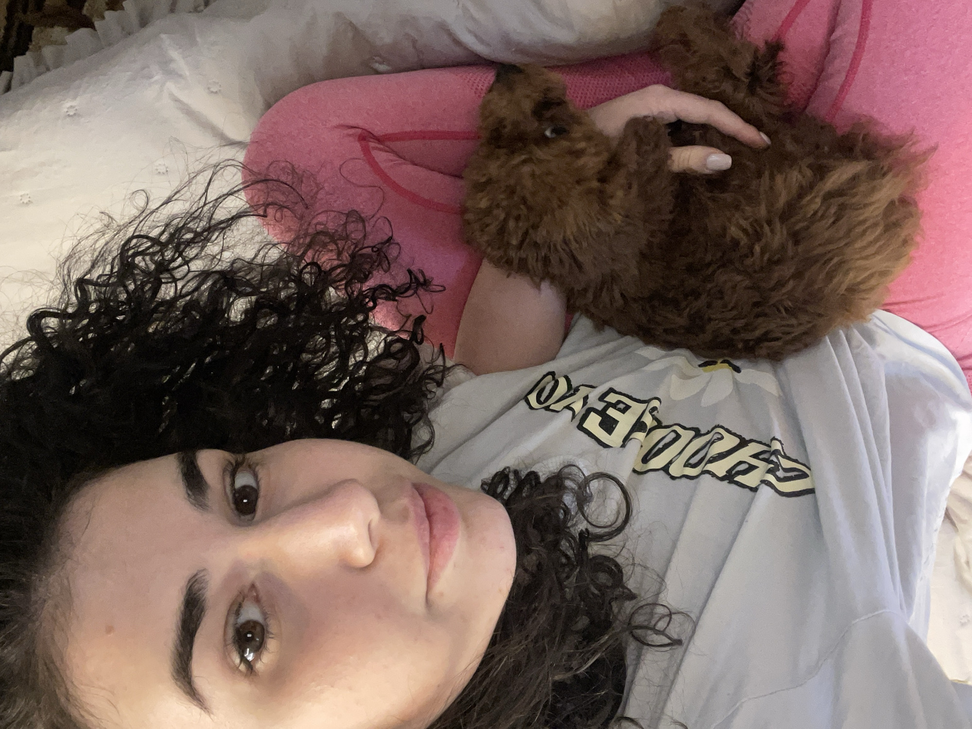 The image depicts an intimate indoor scene in Sofia, Bulgaria, featuring a young woman and a small dog resting comfortably together, likely on a bed. The woman is positioned on her back, looking towards the viewer with a calm and gentle expression. She has dark, voluminous curly hair that fans out around her head on a white, subtly textured pillow or bedding. Her eyes are dark brown, and her features are soft.
She is wearing a light blue t-shirt with a prominent graphic design. The visible text on her shirt appears to be stylized, yellow lettering with a black outline, spelling out "DAISIES," accompanied by a small yellow flower graphic above it. Beneath the t-shirt, a bright pink long-sleeved top is visible on her arms and torso.
Cuddled closely beside her is a small, brown, curly-haired dog, resembling a poodle or doodle breed. The dog is nestled against her side, with its head visible near her shoulder and its body partially resting on her arm. The woman's hand is gently placed on the dog's back, indicating a bond of affection. The dog appears relaxed and content in its position. The bedding beneath them is white, and a pink surface, possibly another pillow or blanket, is also visible. The lighting is soft and ambient, suggesting a cozy indoor environment without specific time-of-day cues. The overall mood of the image is peaceful and warm, highlighting the close companionship between the woman and her pet.