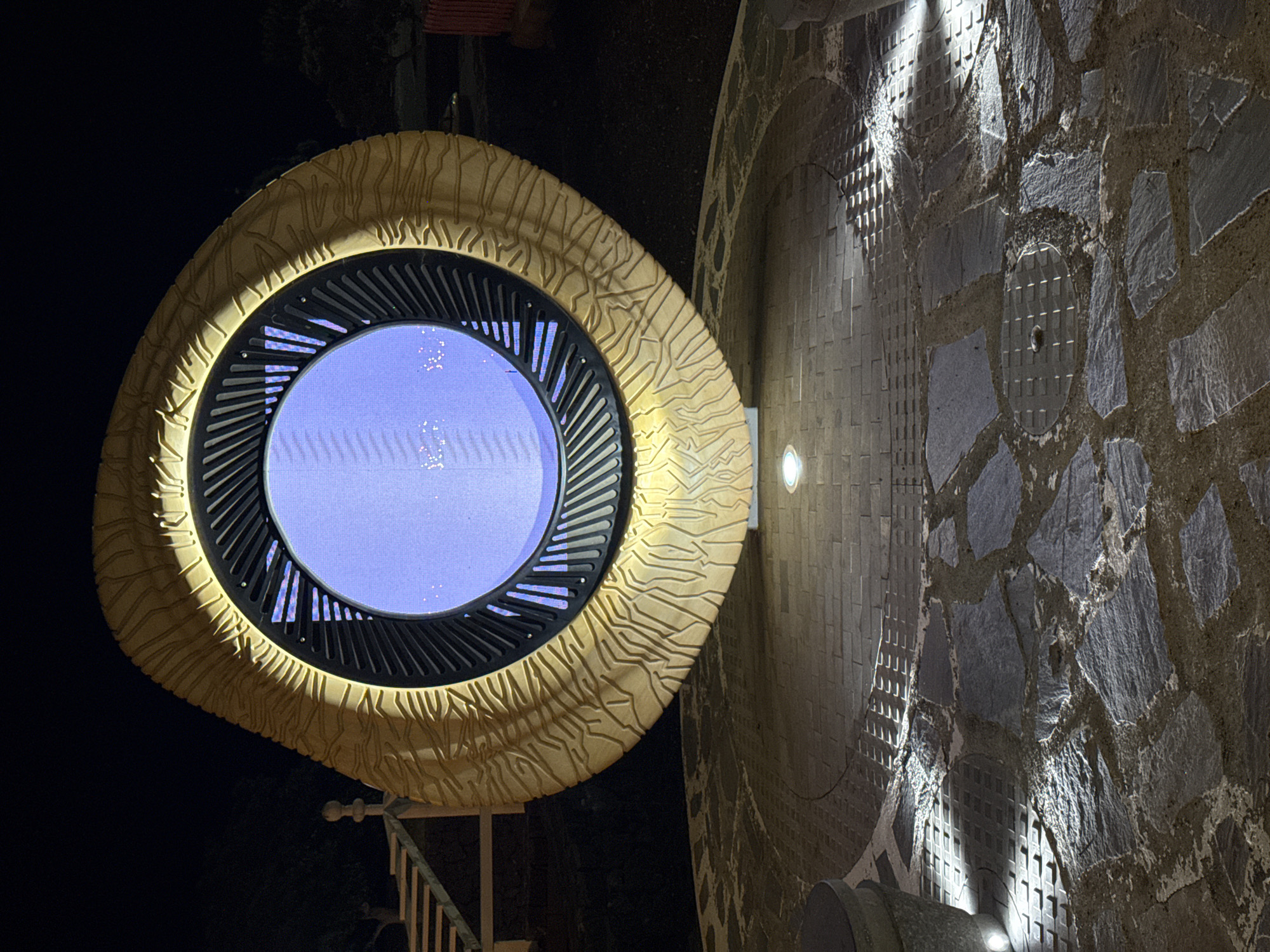 Captured at night, the image showcases an artistic architectural detail in Tías, Spain. A circular structure, possibly an illuminated window or art installation, features a distinctive beige frame with a textured, organic pattern. The frame surrounds a screen displaying a blue light, with faint patterns suggestive of an image.
The circular element is mounted on a wall adorned with a mosaic of irregular stones, illuminated by spotlights. The overall aesthetic blends modern design with natural materials, creating a visually striking juxtaposition. The rest of the scene is shrouded in darkness.