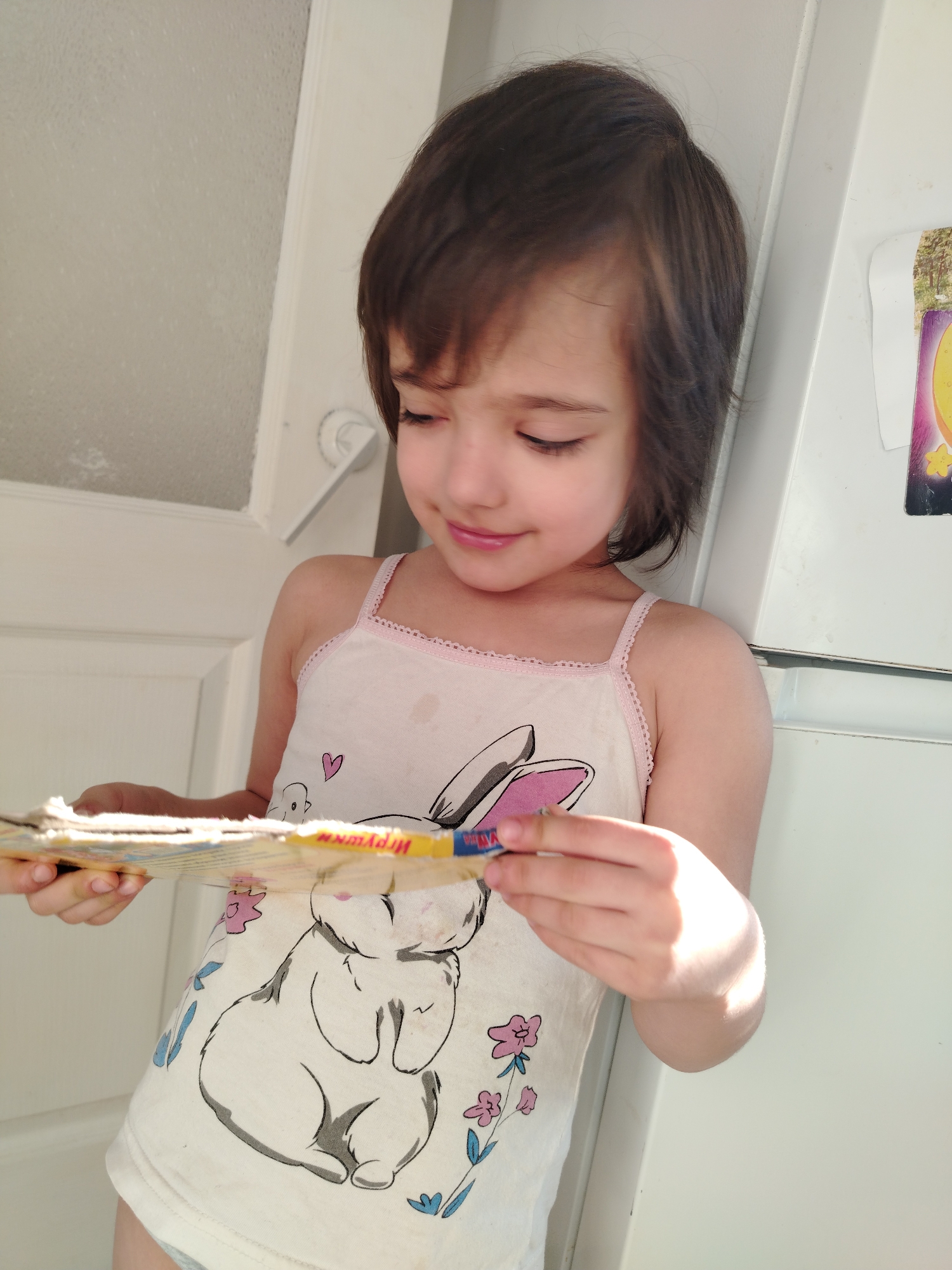 A brightly lit, close-up indoor shot features a young child, likely a girl, from the chest up, engaged in looking at a book or magazine. The child has short, dark brown hair and a gentle, concentrated expression, with a slight smile, as she gazes downwards at the item in her hands. She is wearing a light-colored tank top with thin pink straps and pink lace trim along the neckline. The top is decorated with a large outline drawing of a rabbit with pink ears, a small pink heart, and scattered pink and blue floral patterns.
The child holds a small, somewhat worn book or pamphlet. Visible Cyrillic text on its spine or cover includes "Игрушк" (Igrushk), which likely forms part of the word "toy" in Russian.
The background shows interior elements. To the left, a white door with a frosted glass panel and a white doorknob is partially visible. To the right, a section of a white household appliance, possibly a refrigerator, is seen, with a colorful sticker or magnet affixed to its upper right side. The lighting is bright, suggesting natural daylight, creating highlights on the child's hair, arm, and the book, and contributing to the serene atmosphere. The child appears calm and absorbed in her activity. The setting is indoors, in Noviy, Russia.