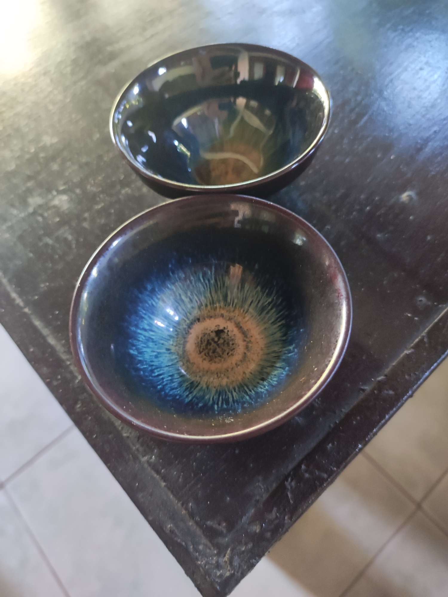 Two small, empty ceramic bowls are positioned on a dark, uneven wooden surface. The closer bowl has a brown ring in the center that transitions to blue and then to a dark brown outer rim. The bowl farther away is primarily dark with reflective surfaces inside. The surface the bowls are on appears to be a corner of a table or other furniture. Part of the floor is visible in the lower right corner, suggesting an indoor location. The absence of people or context points to a staged, product shot likely taken in the Ubud District, Indonesia.