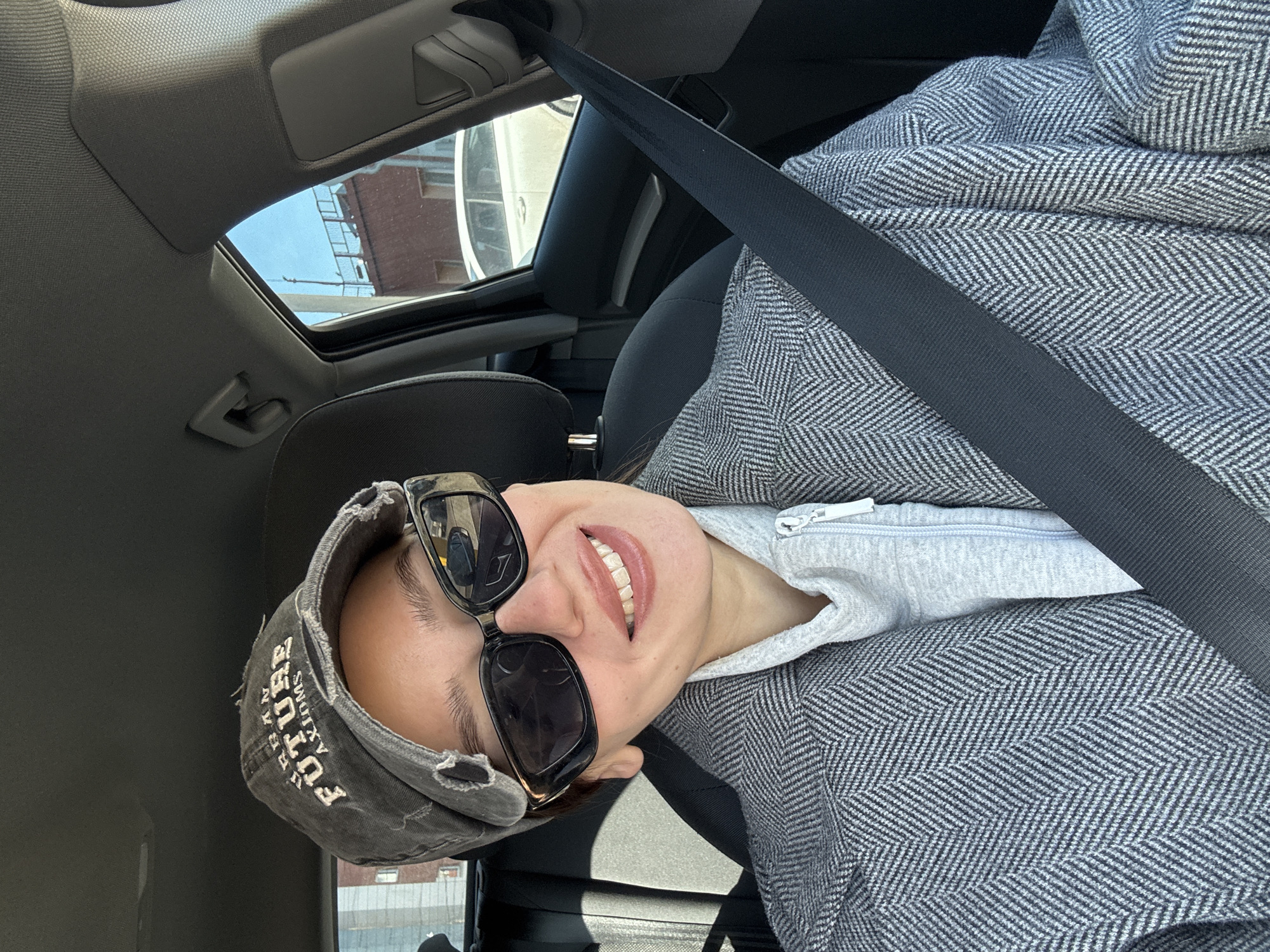 A person is depicted in the front passenger seat of a vehicle during daylight. The individual wears a distressed dark gray baseball cap with "URBAN FUTURE AXIOMS" embroidered in white, and dark rectangular sunglasses. The lower portion of the face is visible, showing a smile with exposed teeth. Clothing consists of a light grey zippered hoodie under a grey herringbone-patterned coat. A black seatbelt is correctly fastened across the chest and lap. The vehicle's interior features dark fabric seating and a light grey headliner.
Through the front passenger window, a reddish-brown, multi-story building with rectangular windows is visible. The building has a flat roof with a dark metal railing and a thin vertical pole or antenna. A white sedan is parked in front of this building. The sky above is clear blue. A lighter-colored building is partially visible through a window on the driver's side.
The scene depicts a generic urban environment with buildings and parked vehicles. The precise location within Krasnoyarsk, Russia, cannot be determined from the visible architectural and vehicle details.