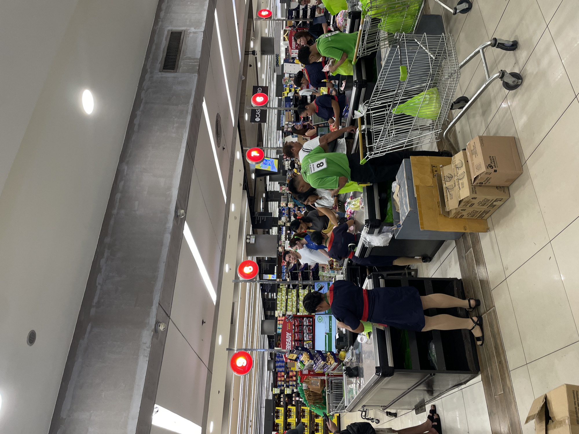 The image shows a busy checkout area in a supermarket, likely in Davao City, Philippines, indicated by the presence of various consumer goods brands commonly found in the region. Several checkout lanes are occupied by customers and cashiers. One cashier, wearing a dark uniform with a red sash, is attending to a customer at the foreground. Another cashier, also in a similar uniform, is seen at a neighboring lane. Several shoppers are waiting in line, some with shopping carts full of groceries. Employees in bright green shirts, some with numbers printed on them (e.g., "8"), are assisting customers, scanning items, and bagging purchases. The environment is brightly lit with overhead fluorescent lights and features typical supermarket elements like shelves stocked with products, cash registers, and shopping carts. The floor is tiled, and a large, grey concrete beam runs across the ceiling.
