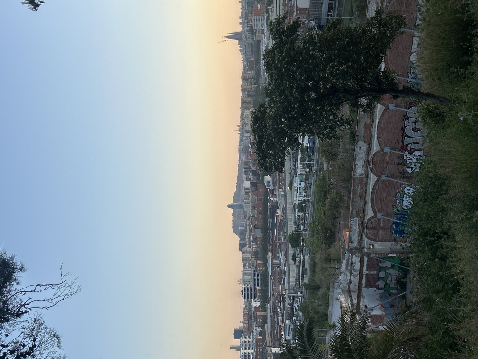 The image shows an elevated view of Santa Coloma de Gramenet, Spain, at dusk or dawn. The sky transitions from blue to orange. The cityscape is densely packed with buildings, including apartment blocks and other structures of varying heights. In the foreground, vegetation such as trees and shrubs frame the view, along with a dilapidated brick structure covered in graffiti. A tall church spire or tower is visible in the middle distance, adding to the skyline. No people are visible in the frame.