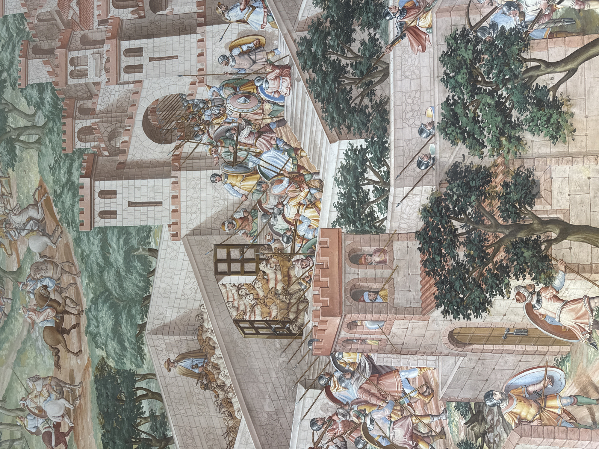 This fresco depicts a bustling scene of military activity and civilian life within and around a fortified city, likely San Lorenzo de El Escorial, Spain, given its characteristic architectural style. The fresco showcases multiple levels and elements.
In the upper portion, horsemen in historical attire are depicted moving across a landscape with trees. Below them, the city walls and towers are prominent. A large gatehouse with an arched entrance is a focal point, with many soldiers armed with spears and shields gathered both inside and outside the gateway. Some figures are seen climbing stairs towards the upper ramparts, indicating movement and defensive preparations.
On the left side of the fresco, a shepherd with a staff guides a flock of sheep up a stone staircase towards a fortified area. Further down on the left, a soldier in a yellow tunic and a blue shield stands guard.
Throughout the scene, numerous individuals are depicted in various states of action. Soldiers are shown in formation, some with helmets and armor, others with spears and shields. Civilians, including the shepherd and people interacting near the gate, are also visible. The architecture is detailed, featuring stone blocks, battlements, and arched openings. Trees with dense green foliage are interspersed throughout the scene, providing a natural contrast to the man-made structures. The overall impression is one of a dynamic and well-defended settlement.