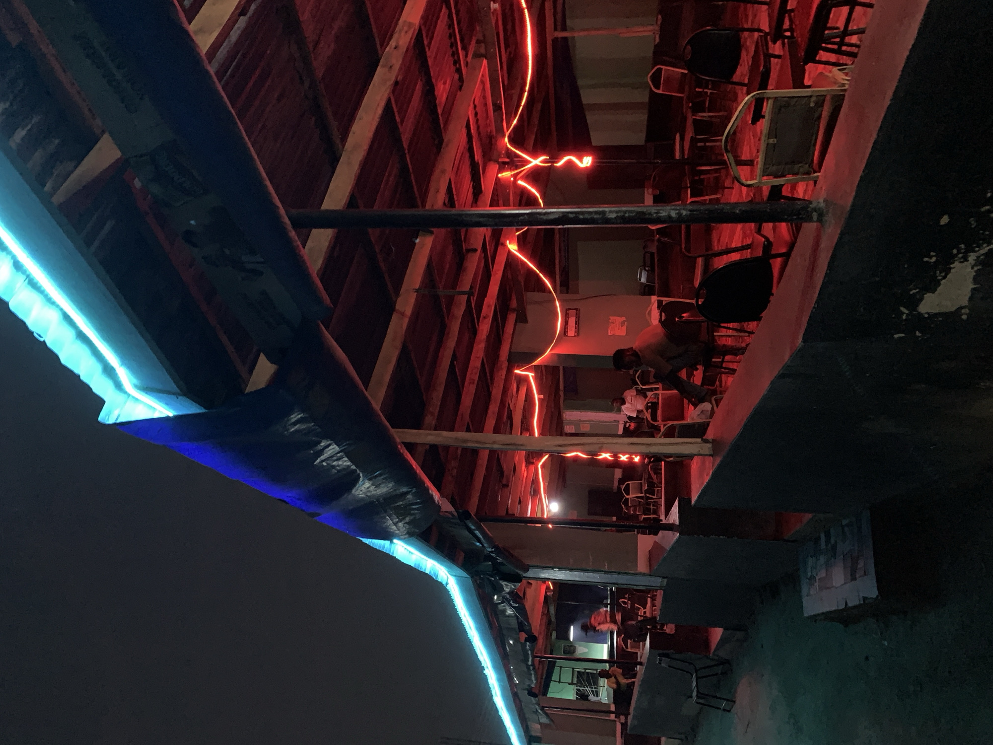 The image depicts an outdoor setting in Kabong, Nigeria, at night. The architecture consists of a wooden structure, possibly a restaurant or bar, illuminated by vibrant blue and red neon lights. Several people are seated at tables and chairs within the structure. The surrounding environment appears dimly lit with a mix of urban elements.
