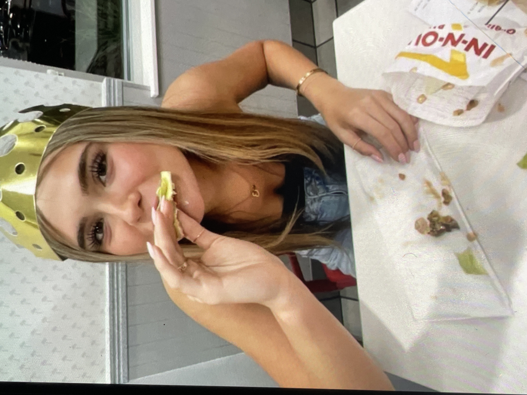 A woman with long blonde hair and a gold crown on her head is eating food from a partially eaten In-N-Out Burger bag. The bag and remnants of food are on a white surface. The woman is wearing a black top and denim jacket. She has a gold bracelet and necklace. The location is not Harmanli, Bulgaria.