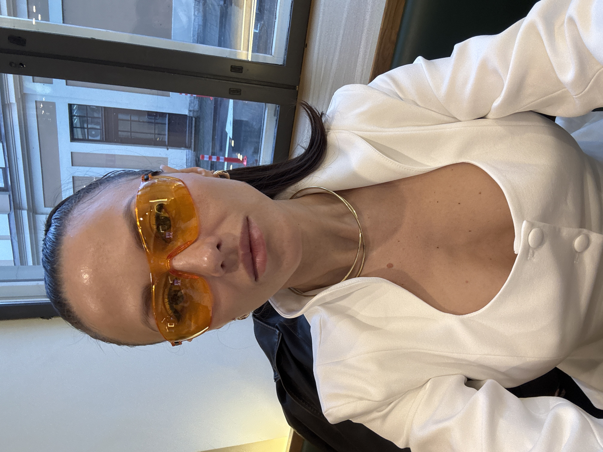 A woman with dark hair pulled back and wearing distinctive orange sunglasses is seated near a window in what appears to be a building in Saint Petersburg, Russia. Outside the window, architectural details of a building can be seen. The woman is wearing a white top or jacket with buttons near the neckline and a gold-colored necklace. A black garment, possibly a jacket, is visible behind the white top. The lighting suggests it might be daytime or early evening.