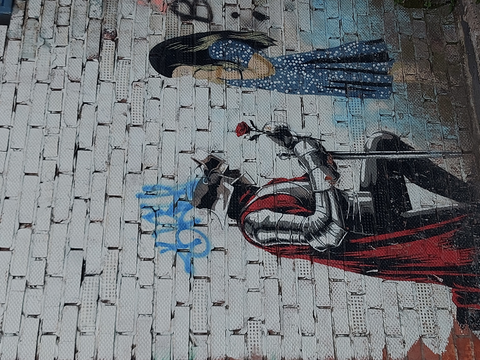 This is a photograph of a graffiti artwork located in the city of Izhevsk, Russia. The artwork depicts two primary figures painted on a wall constructed of small, white, brick-like blocks.
The upper figure is a young woman with dark hair, lying down, and wearing a blue dress with white polka dots. The lower figure is a knight on horseback. The knight is in silver armor with a red cloak. The knight is holding a red rose. There is blue graffiti near the knight's head.