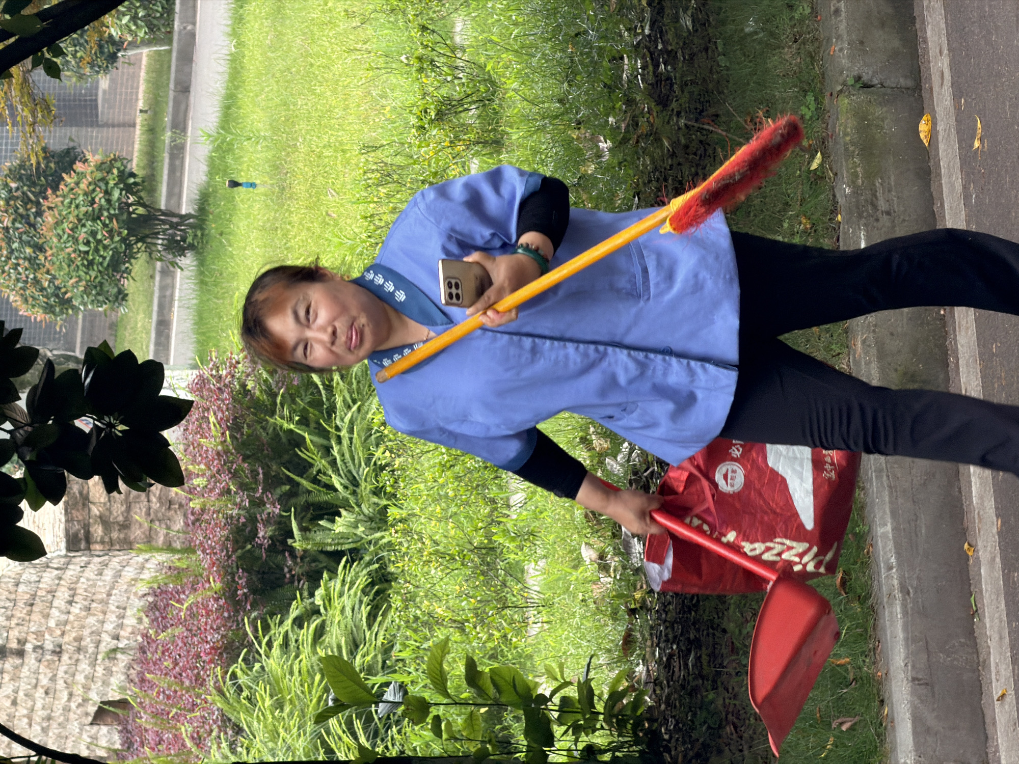 A middle-aged Asian woman is walking outdoors in what appears to be a park or street in Jiangjin, China. She is wearing a light blue collared uniform with dark blue trim around the collar, black undersleeves, and black pants. She is holding a yellow pole with a red bristled duster or brush attachment in her right hand, and a red dustpan in her left hand. She is also carrying a large red reusable shopping bag with white lettering, which includes the word "Pizza" and Chinese characters. In her right hand, she is holding a gold-colored smartphone. The woman is looking towards the camera and has a slight smile. The background consists of lush green foliage, including ferns and bushes with red leaves, a stone building or wall, and a paved area with a concrete curb. There are fallen leaves on the ground.
