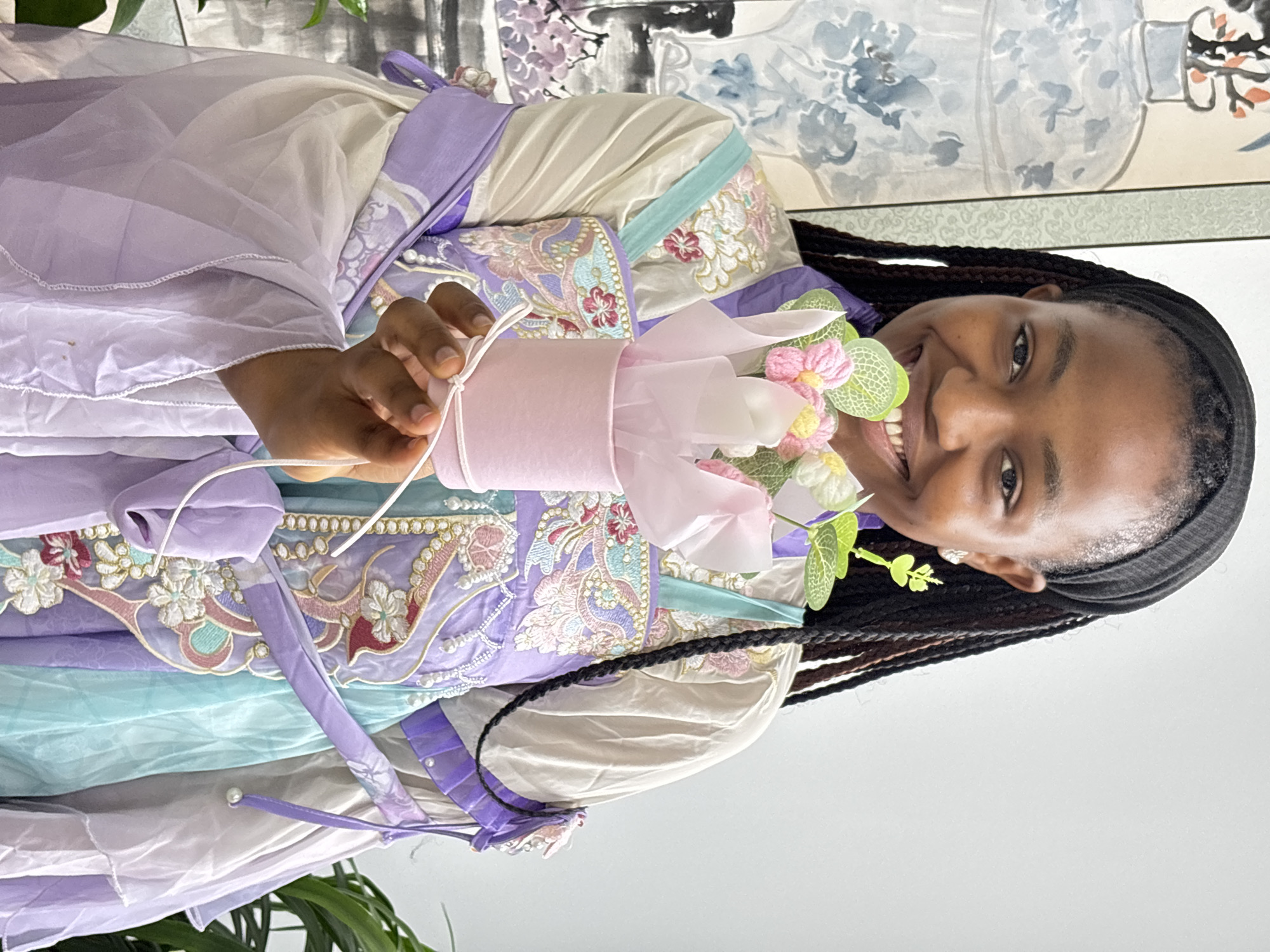 A young woman with dark braided hair smiles at the camera while holding a small pink container filled with artificial flowers. She is wearing a traditional garment with layers of lavender, cream, and light blue fabric adorned with floral embroidery. The background includes a wall with a traditional Chinese painting of flowers. Jiangjin, China is not present in the image.