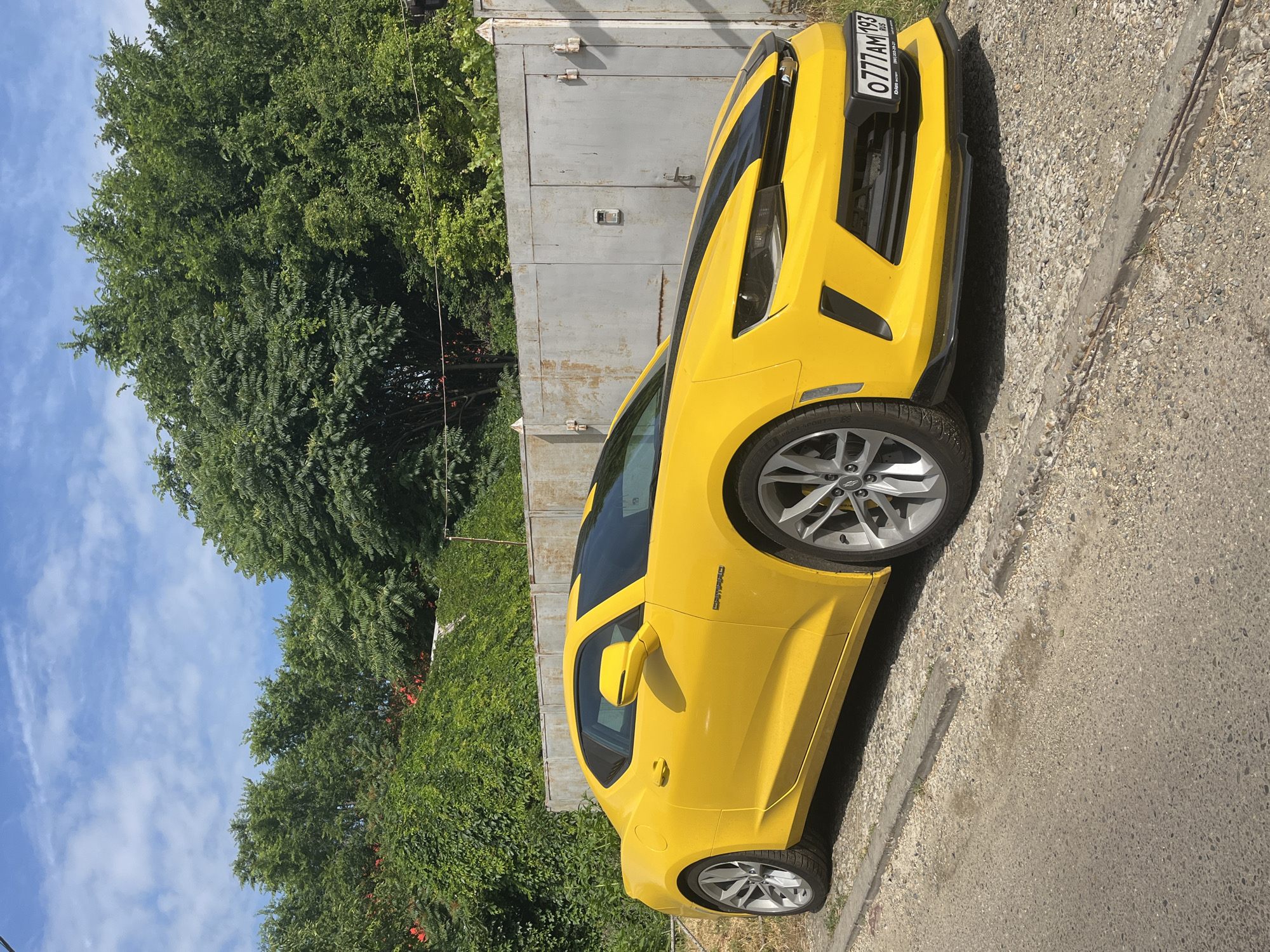 A bright yellow Chevrolet Camaro is parked on a gravel shoulder next to a paved road. The car has black racing stripes on the hood and roof. Behind the car is a weathered, corrugated metal garage door and lush green trees. A license plate on the front of the car reads "0777 AM 193 RUS". The sky is blue with scattered white clouds. The scene appears to be in a residential or industrial area of Krasnodar, Russia, given the license plate format and general landscape. There are no people visible in the image.