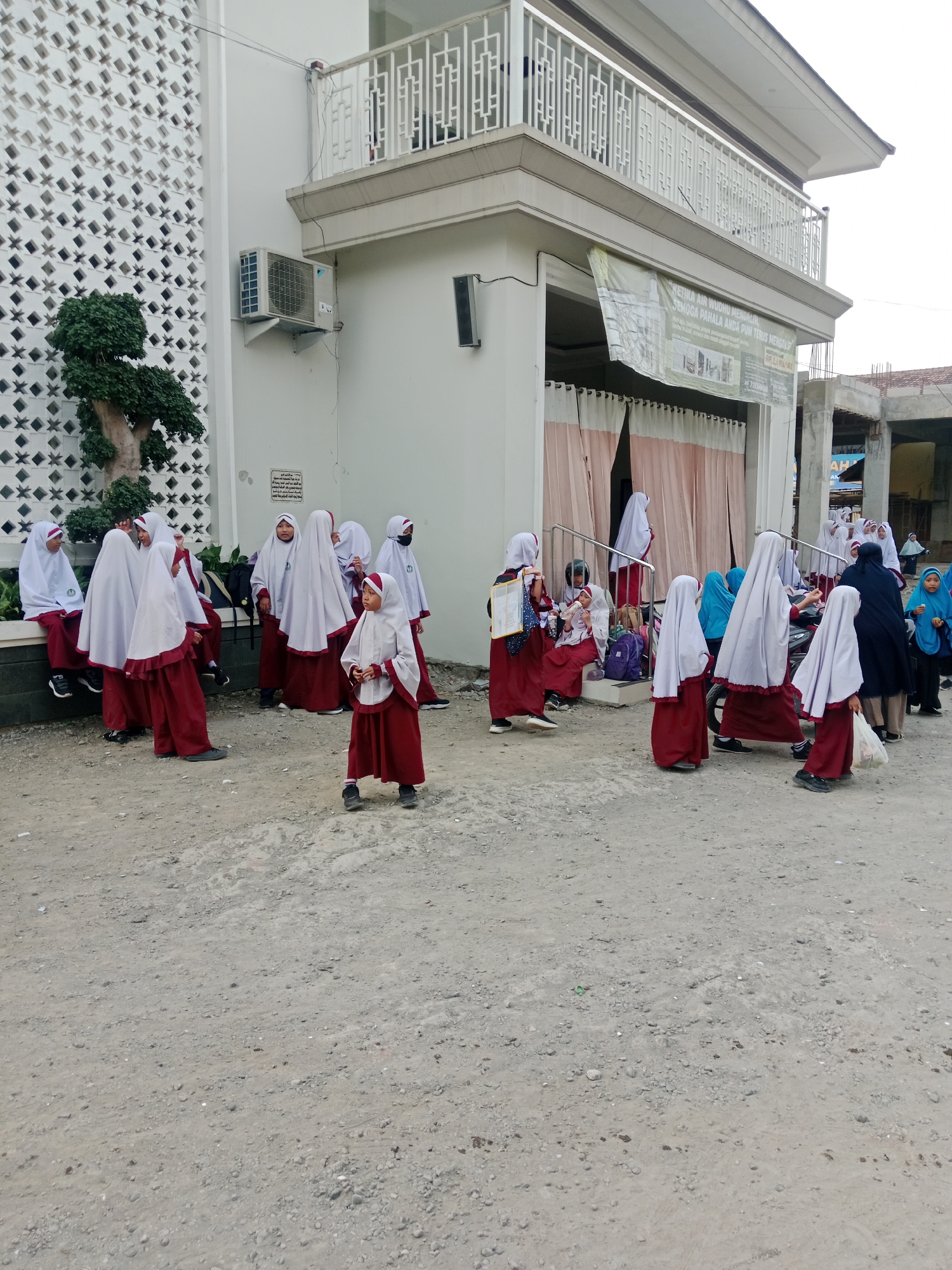 A group of young girls wearing maroon and white Islamic school uniforms are gathered outside a building with a white facade, possibly a mosque or school, in Kediri City, Indonesia. The building features a balcony with decorative railings and an air conditioning unit mounted on the exterior wall. Several of the girls are in motion, while others are seated or standing in conversation. Some girls are wearing hijabs that are predominantly white, while others are wearing hijabs that are a blend of white and maroon. The ground is a rough, dirt surface with some scattered stones. A banner is hanging from the building, partially visible and displaying text. To the right, a construction site is visible with scaffolding and incomplete structures, suggesting ongoing development in the area. The lighting suggests it is daytime, possibly overcast.