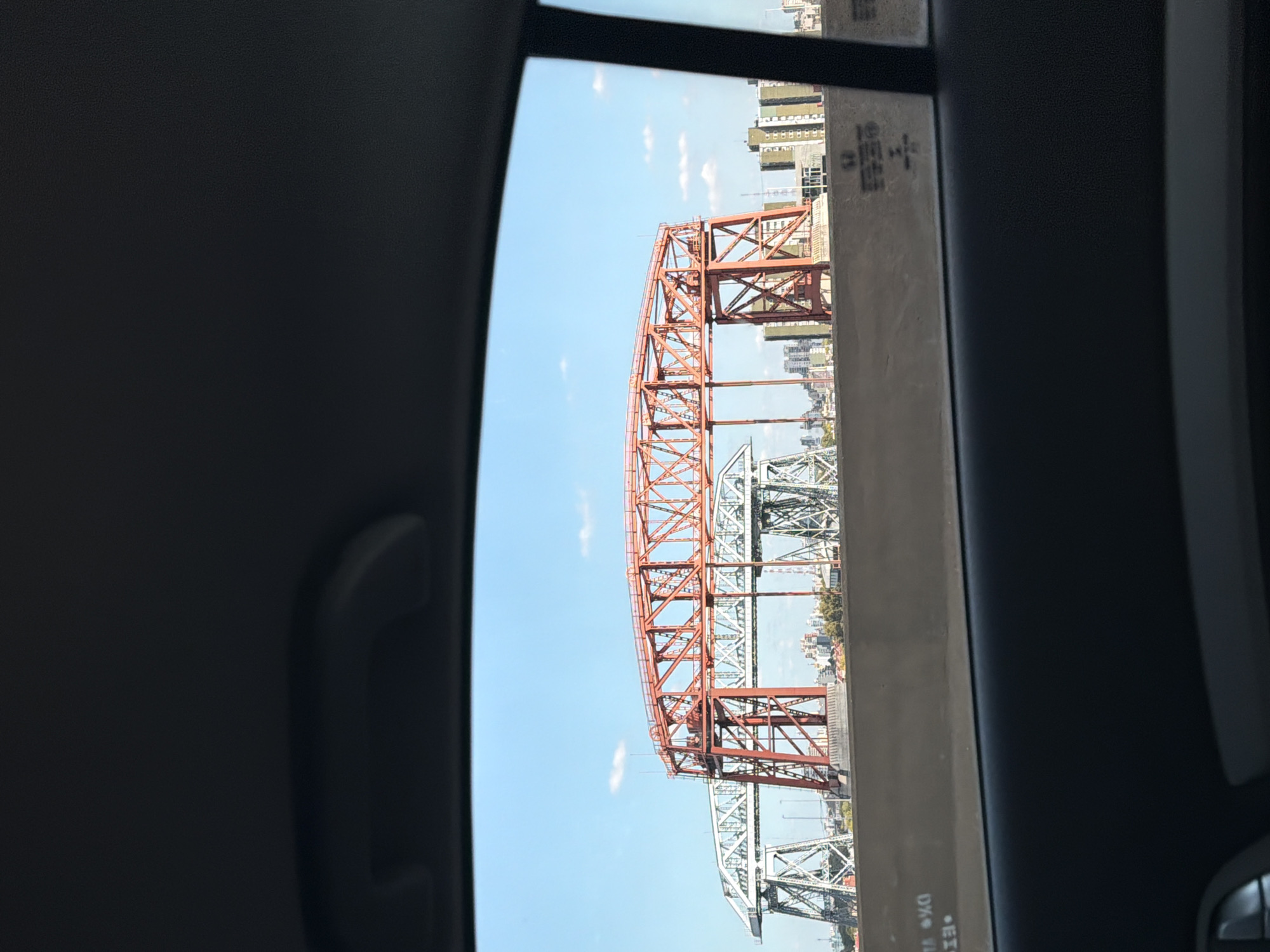 The image shows the Nicolás Avellaneda Bridge in Buenos Aires, Argentina, as seen from inside a vehicle. The bridge is a red and green steel structure spanning water, with buildings visible in the background. The view is framed by the vehicle's interior, including a window and door panel. The sky is blue with a few clouds. There are no people visible in the frame.