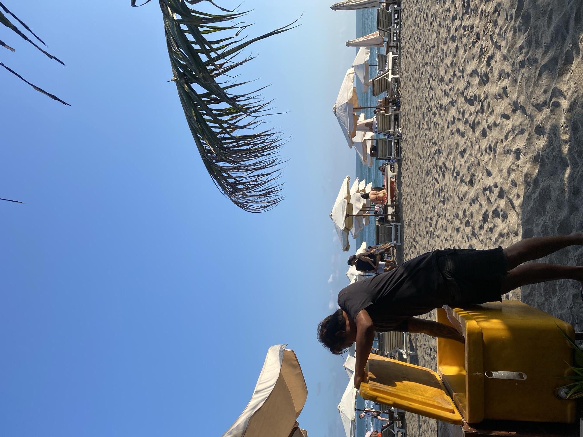 A man wearing a black t-shirt and black shorts is standing next to a large, yellow cooler on a sandy beach in North Kuta, Indonesia. He is leaning over the open cooler, reaching inside with his right hand. The beach is lined with white lounge chairs and umbrellas. In the background, the ocean is visible with gentle waves and a clear blue sky. Several people are scattered on the beach, some relaxing on the lounge chairs and others walking near the water. Palm fronds are hanging into the frame from the top right.