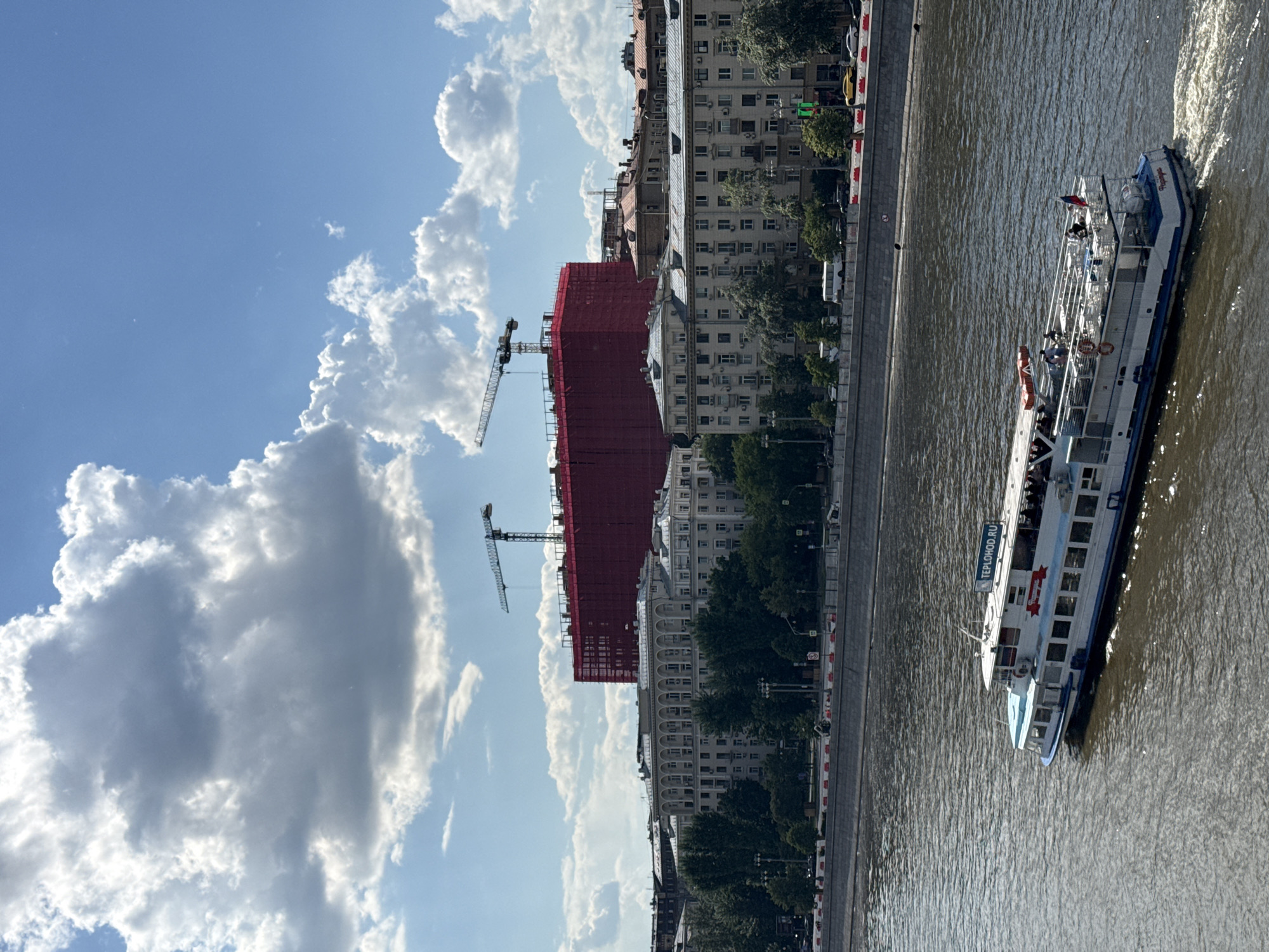 The image depicts a vibrant daytime scene along an urban river, likely the Moscow River, under a bright blue sky dotted with large, fluffy white cumulus clouds.
In the foreground, a white and blue passenger boat, identified by the text "TEPLOHOD.RU" and a red star emblem on its side, cruises along the brownish water. The boat is moving towards the right, leaving a distinct white wake behind it. Several individuals are visible on the boat's upper deck and through its windows, indicating a leisure or sightseeing activity. A small Russian flag flies from the stern.
The far bank of the river features a tree-lined embankment with a paved road or promenade, where a few vehicles and people are sparsely visible. Behind this, a dense urban landscape unfolds. A large building under construction is prominent in the midground, entirely enveloped in red safety netting, with two tall tower cranes extending upwards above it. Adjacent to and beyond the construction site are numerous multi-story buildings, consistent with older urban residential or commercial architecture.
The bright sunlight suggests it is daytime, possibly mid-morning or afternoon, with clear weather conditions apart from the scattered clouds. The scene conveys a sense of active urban life and recreation on the water.