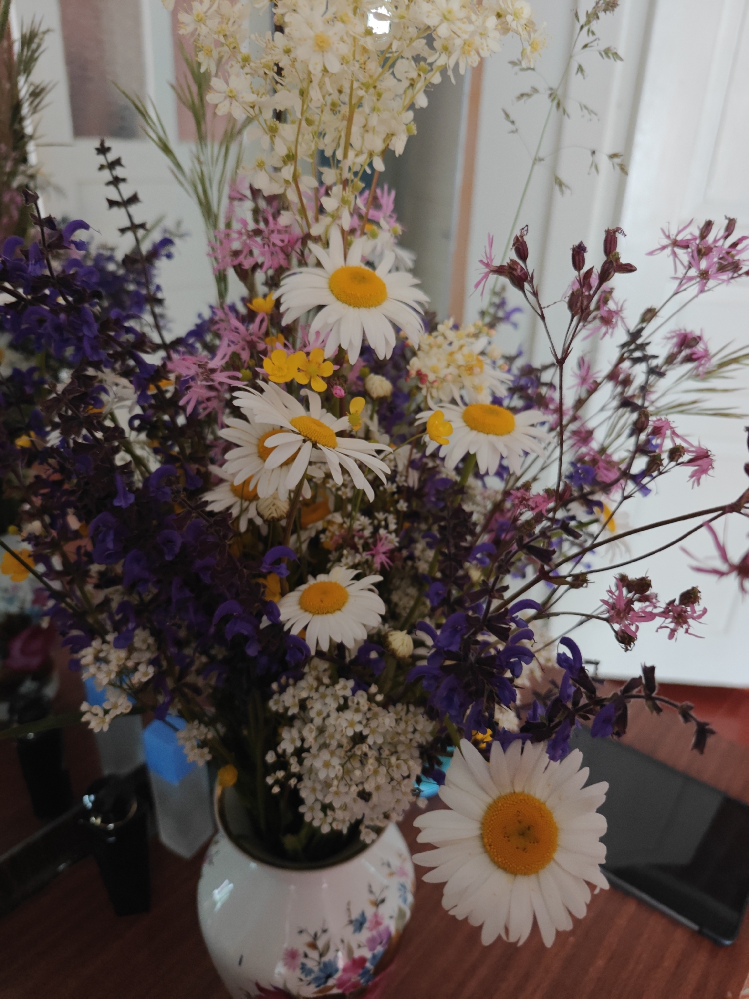 A close-up shot of a vase filled with various wildflowers. The arrangement includes white daisies with yellow centers, purple flowers, small white blossoms, and pink spindly flowers. The vase is white with a floral pattern. The table is a dark reddish-brown color. A smartphone is on the table next to the vase, along with a black cup. The background appears to be a mirror. The location is Knyshivka, Ukraine.