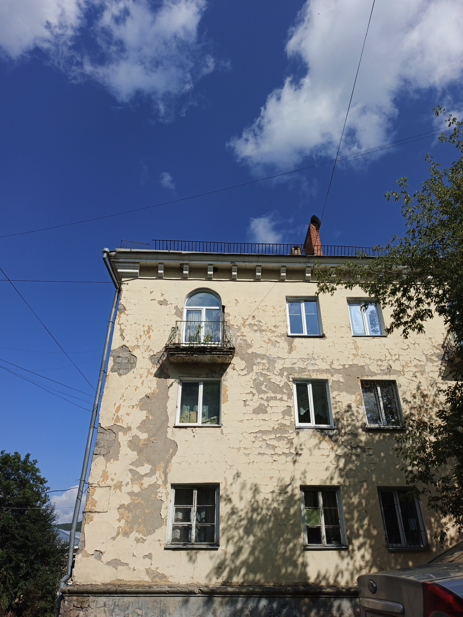 The image depicts the facade of an old, weathered apartment building in Zlatoust, Russia, under a bright blue sky with scattered clouds. The building's exterior plaster is peeling extensively, revealing underlying brickwork in places. The building has multiple windows, some with white frames and others with older, darker frames. One window on the second floor is arched and features a small balcony with a black metal railing, holding potted plants. A chimney made of red brick is visible on the rooftop, which also has a black metal railing. Electrical wires crisscross the sky above the building. A large tree with green leaves casts shadows across the building's facade. The lower part of the building appears to be made of stone. In the foreground, the rear of a car is partially visible. The scene is typical of an urban residential area.