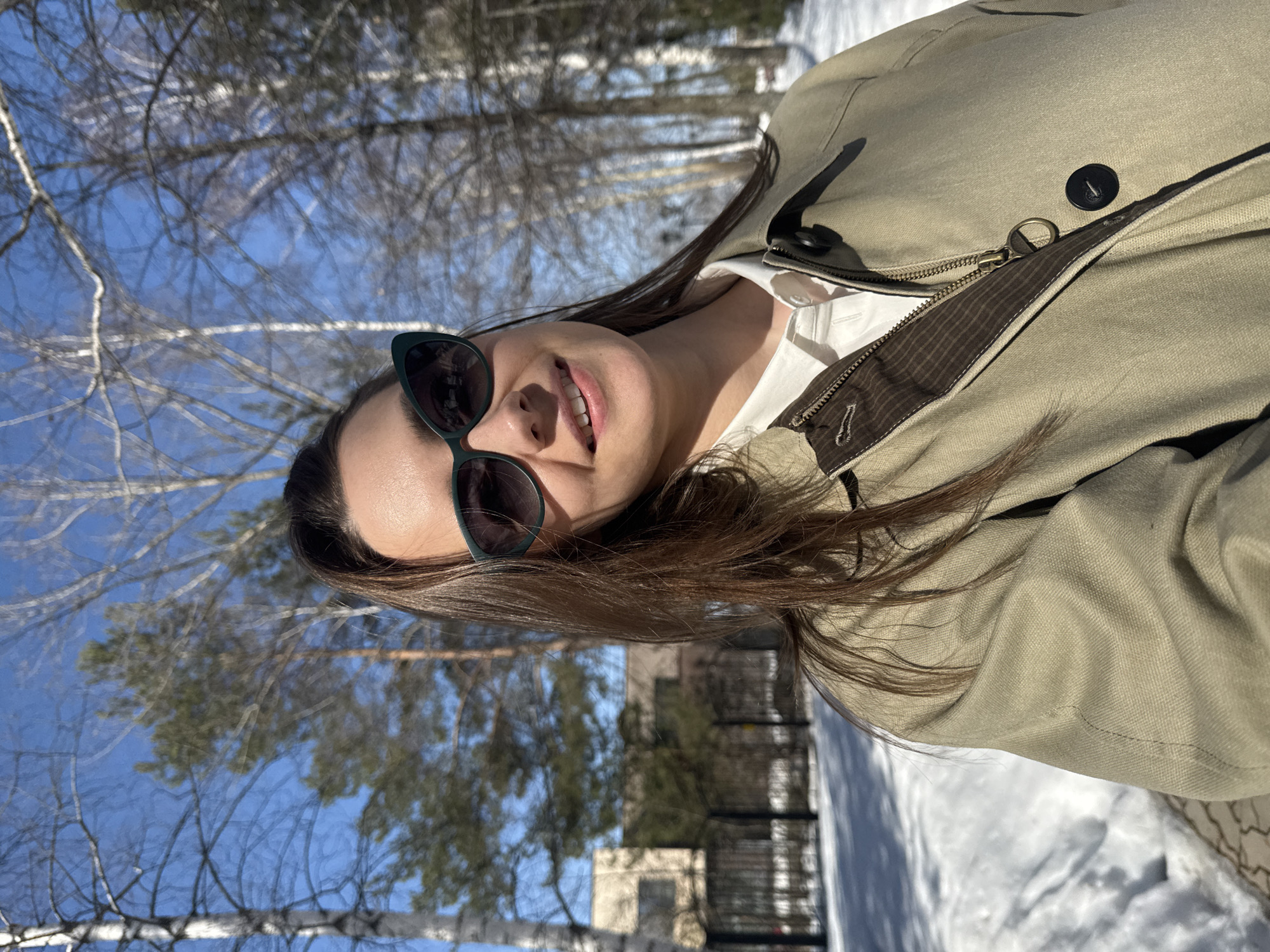 A woman is posing for a selfie outdoors in Chaikovsky, Russia, with trees in the background and snow on the ground. The woman has long, dark hair, sunglasses, and is wearing a light green jacket over a white shirt. The background features bare trees against a bright blue sky. Evidence of buildings and perhaps other people are present, though blurred or distant. The season is likely late winter or early spring, given the snow and lack of leaves on the trees.