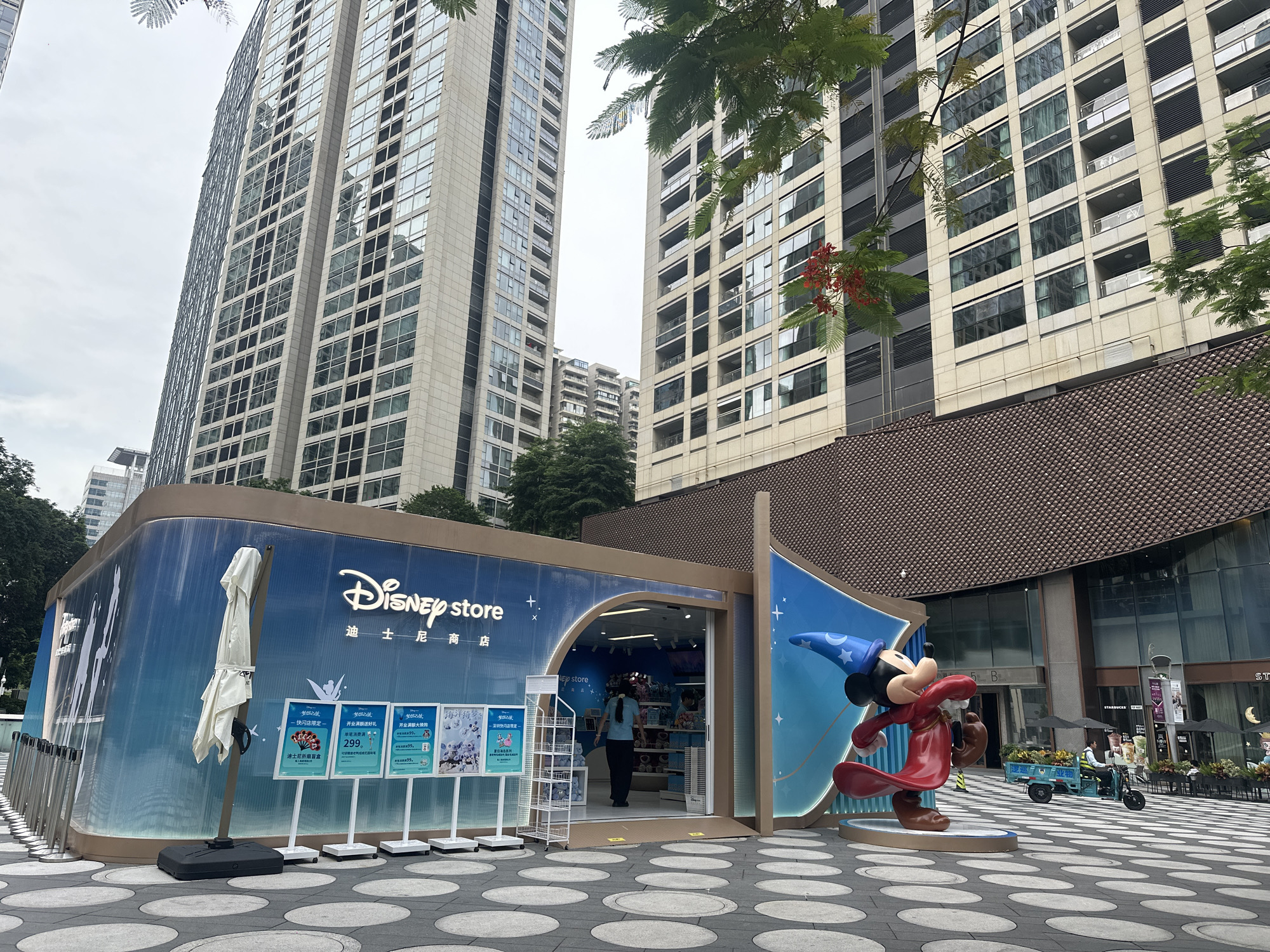 The image depicts a temporary Disney Store pop-up shop located in Futian District, China. The store features a bright blue facade with the Disney logo prominently displayed, along with a large statue of Mickey Mouse in his Sorcerer Mickey costume. The store appears to be open, with merchandise visible inside and a woman standing at the entrance. In the foreground, a checkered pavement with circular inlays surrounds the store. Behind the pop-up, tall, modern buildings dominate the cityscape, their glass facades reflecting the overcast sky. A street scene unfolds to the right, with a man operating a small electric vehicle laden with goods, and umbrellas covering outdoor seating areas of adjacent businesses, including what appears to be a Starbucks. The overall impression is one of a bustling urban environment with a touch of whimsical branding.