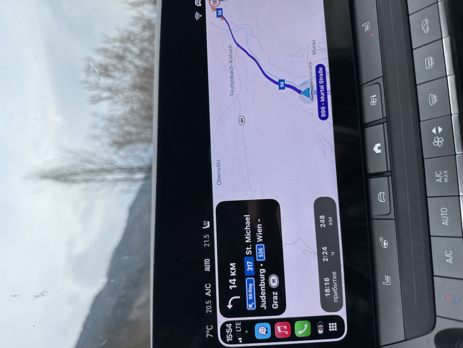 The image presents a forensic view from inside a vehicle, centered on its active infotainment system and dashboard controls, with the outside environment partially visible through the windshield.
**Location:** The navigation map on the infotainment screen clearly indicates the vehicle's current position (marked by a blue triangle) on the "B96 - Murtal Straße," in the immediate vicinity of Murau, Austria. Towns such as Oberwölz and Teufenbach-Katsch are also displayed on the map.
**Objects/Subjects:**
* **Infotainment Screen:** A dark-themed digital display showing a navigation application, likely Apple CarPlay.
* **Top Left Panel:** Displays "7°C" (outside temperature), "20.5 A/C", "AUTO", "21.5" (interior target temperature), and a fan icon. Connectivity indicators "LTE" and signal bars are present.
* **Left Navigation Panel:** Shows a left arrow indicating an upcoming turn in "14 KM". Listed destinations include "RB Ring", "317 St. Michael", "Judenburg • 536 Wien", and "Graz". Apple CarPlay app icons for messages, music, phone, maps, and settings are visible.
* **Bottom Navigation Panel:** Displays "18:18" (estimated arrival time, labeled in Cyrillic), "2:24" (remaining travel time), and "248 KM" (remaining distance).
* **Map Display:** A purple route is highlighted on the map, showing the vehicle on the B96 - Murtal Straße. A speed limit sign of "100" is visible further along the route.
* **Dashboard Controls:** Below the screen, physical buttons are visible:
* **Upper Row:** Controls for heated seats (driver and passenger), car settings, and home screen.
* **Lower Row:** Climate control buttons including "A/C", "AUTO", "A/C MAX", fan speed adjustment, and defroster functions.
* **Exterior View (blurred):** Through the top portion of the windshield, an overcast sky is visible. To the left, a dark, distant mountain range or large hill is present. To the right, blurred bare deciduous trees or shrubs indicate a winter or early spring season. The windshield has minor spots or streaks.
**Actions/Interactions:** The vehicle is either currently in motion or recently was, with the navigation system actively providing directions and the climate control system set to automatic operation. No individuals are present in the frame.