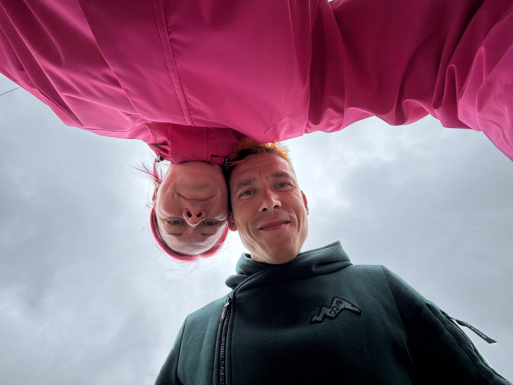 The image is a low-angle selfie of two adults, taken outdoors against a cloudy sky. The person on the left is wearing a dark green hooded jacket with a zipper and a logo on the chest. This person has short, light-colored hair and is smiling. The person on the right is wearing a bright pink jacket. This person has red hair and is also smiling, with a nose ring visible. They appear to be leaning in towards the camera, close together. The overcast sky suggests a cool or possibly damp day. The location is stated to be in the city of Chaikovsky, Russia.