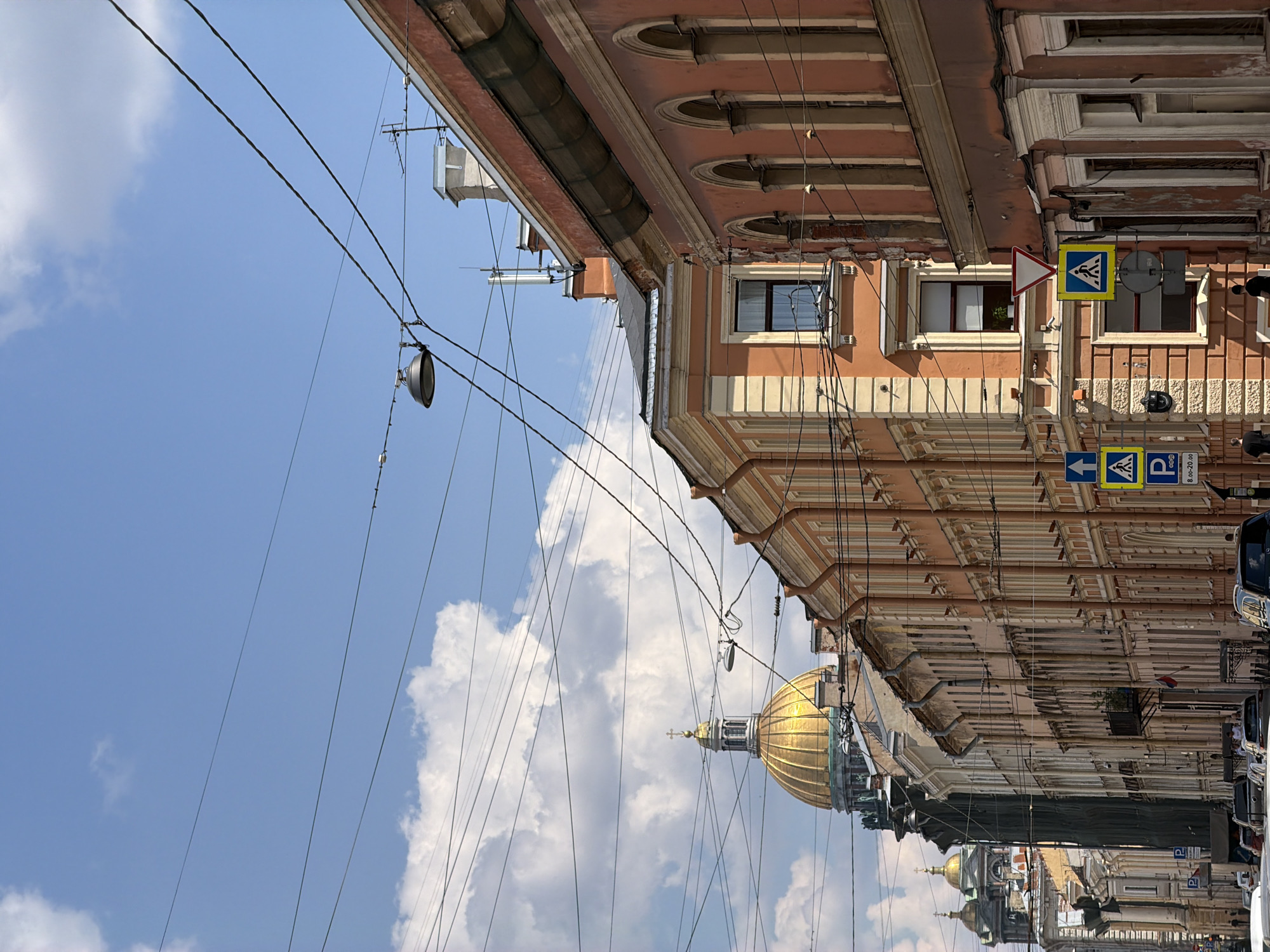 The image presents a vibrant street scene in Saint Petersburg, Russia, under a bright blue sky dotted with scattered white clouds. Ornate, multi-story buildings with peachy-orange facades and intricate white architectural details, including arched windows and cornices, line the right side of the frame.
Numerous black utility wires crisscross the sky, some suspending streetlights or lanterns. In the mid-ground, a prominent golden dome with a cross, characteristic of St. Isaac's Cathedral, rises above the rooftops, with smaller golden domes visible further down the street. Parked cars are partially visible at the bottom of the frame.
The time of day appears to be sunny morning or afternoon, with clear weather. Visible road signs include yellow square pedestrian crossing signs, a red triangular "give way" sign, and blue rectangular parking signs. One parking sign displays "P" and "8.00-20.00", indicating parking hours. A small Russian flag is also visible on one of the buildings. The scene captures the bustling, historical urban environment of Saint Petersburg.
