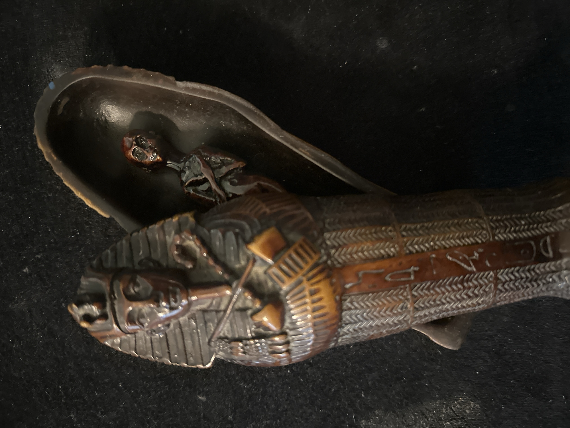 The image shows a dark brown, carved object resembling an Egyptian sarcophagus. The sarcophagus is intricately detailed with hieroglyph-like carvings. Inside, a small, skeletal figure is visible, partially emerging from a dark, boat-shaped vessel. The object is placed on a black, textured surface, likely fabric. The lighting creates strong shadows, highlighting the texture and contours of the carving. No specific location in Togliatti, Russia is depicted or suggested by the image.