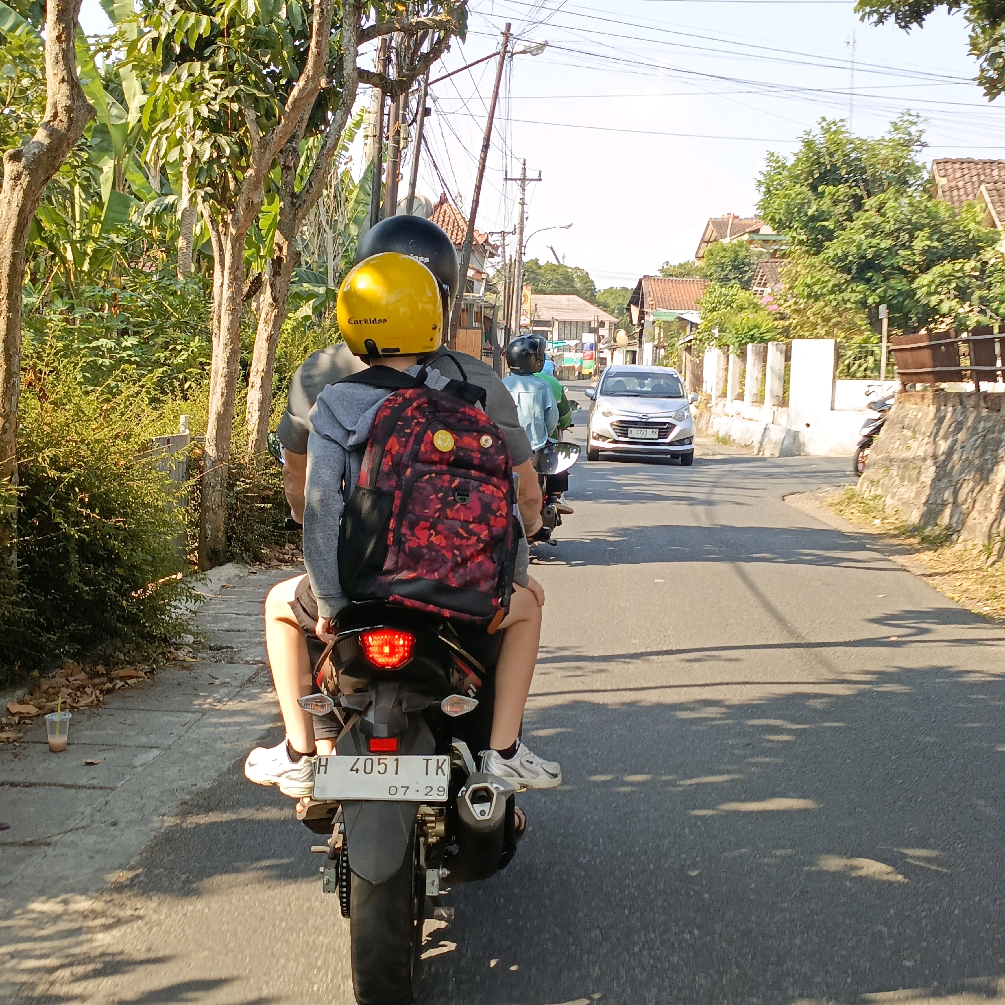This image depicts a street scene in Salatiga, Indonesia, likely during daylight hours. In the foreground, two individuals are riding a motorcycle. The person in the back is wearing a yellow helmet with the brand "CarKidss" visible and a black and red patterned backpack. The person in the front is wearing a black helmet. They are moving away from the camera, down a paved road lined with trees and vegetation on the left, and buildings on the right. A silver car is visible further down the road, also moving in the same direction. The environment suggests a residential or semi-rural area with overhead power lines present. The sun is casting shadows on the road, indicating a sunny day.