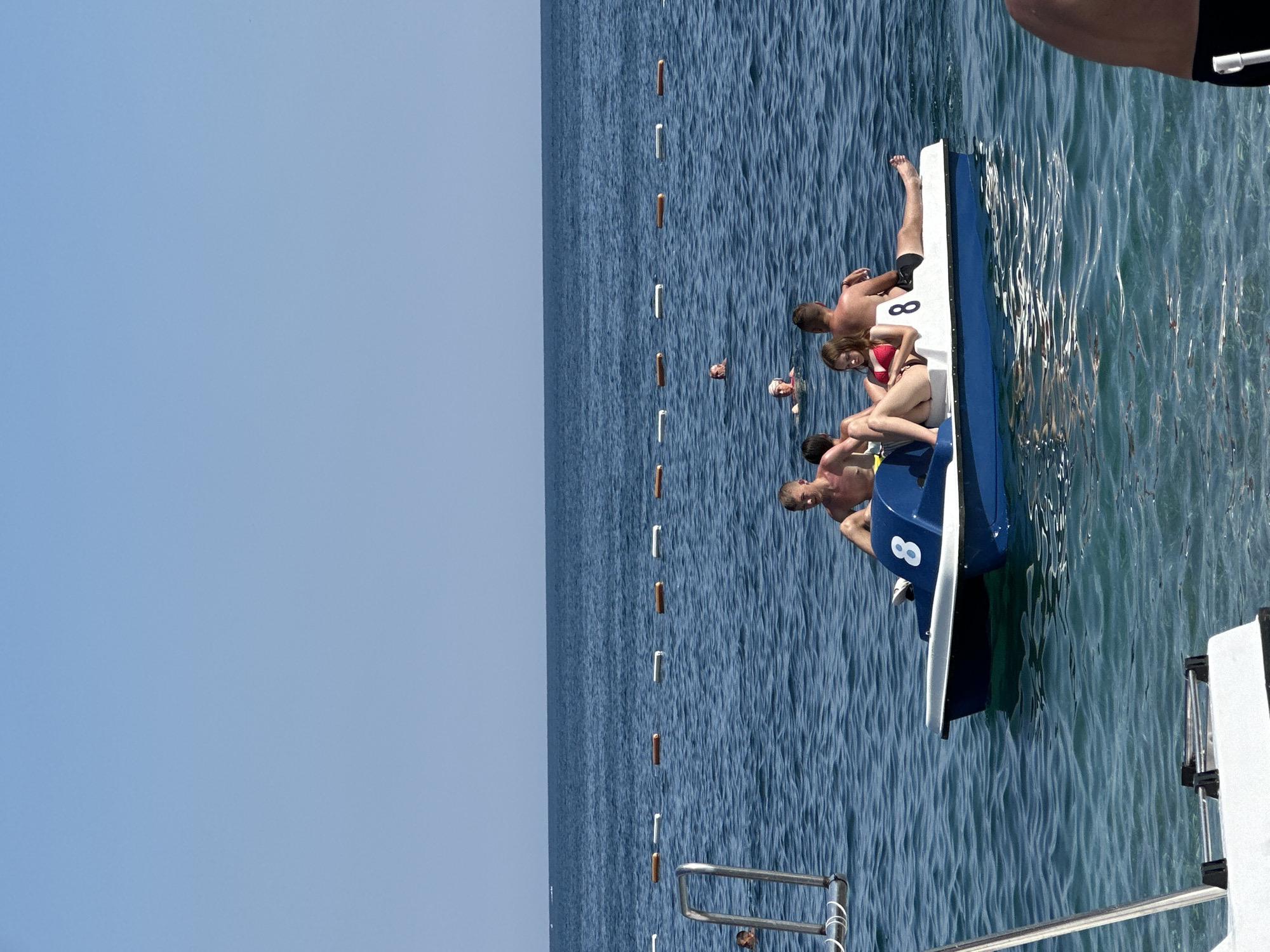 In this outdoor, daytime shot, a group of young adults are enjoying a day on the water near Budva, Montenegro. The scene is dominated by the calm, blue sea, with a clear, bright sky above. A few people are swimming in the water, while others are seated on a small, white and blue pedal boat.
On the pedal boat, three individuals are visible. One person, wearing black shorts, sits with their legs extended over the side of the boat. Next to them, a woman in a red bikini is seated, looking towards the viewer. Another person, with their back to the camera, is leaning over the edge of the boat. A number "8" is clearly visible in white on the side of the blue portion of the pedal boat.
Other swimmers are scattered in the water. One person's head is visible just above the surface, and another appears to be further out in the sea. The water has a rippling texture, suggesting a gentle breeze. There are several orange and white buoys lined up in the water, possibly marking a swimming area or a course. In the foreground, a metal railing is visible, likely part of a larger structure or another boat. The overall mood appears relaxed and recreational, consistent with a summer day at the beach.