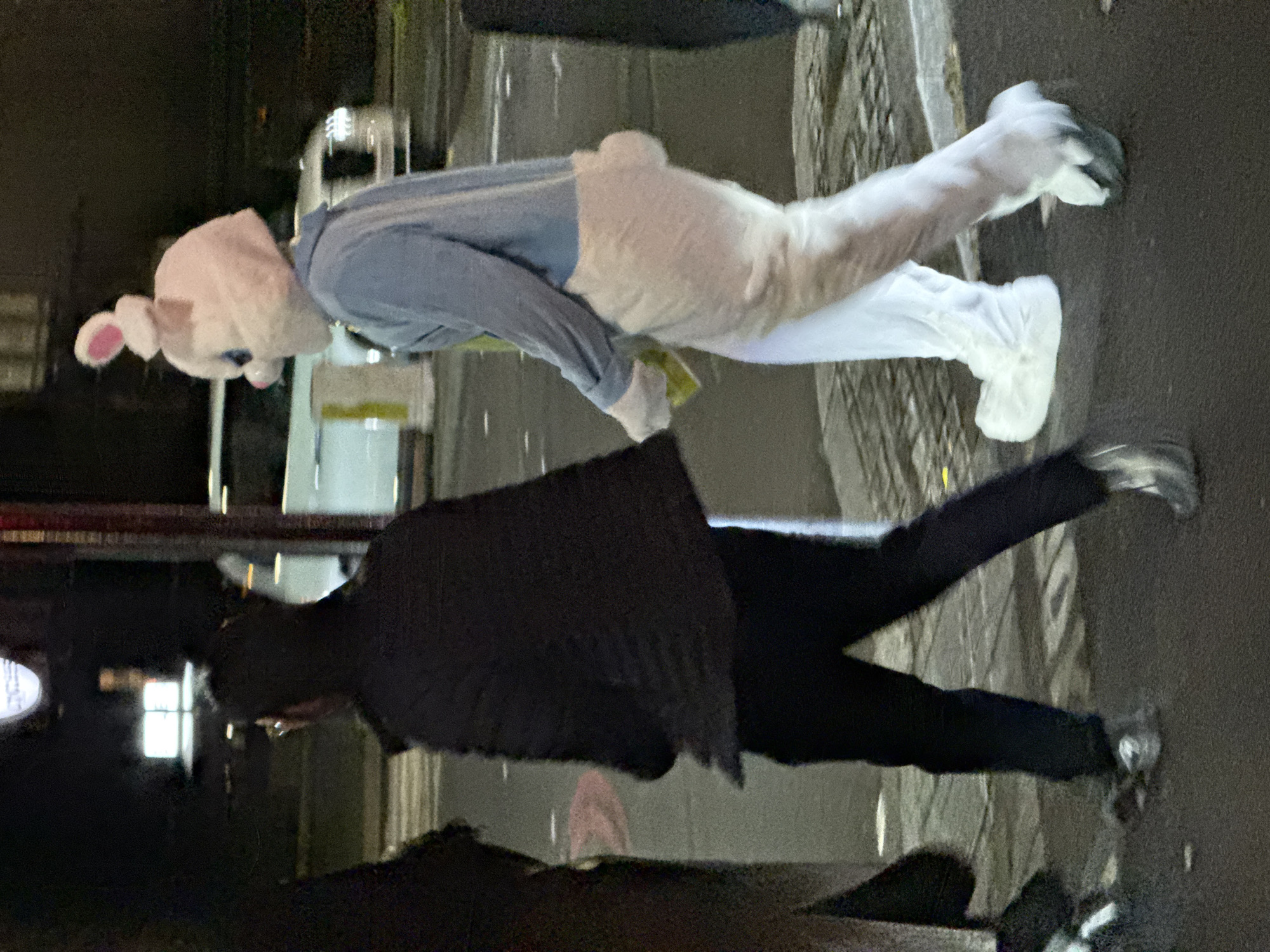 The image shows two individuals on a street. One individual is wearing a bunny costume with a pink head, blue shirt, and white trousers. The other person is wearing dark trousers and a dark jacket. The setting appears to be an urban environment at night. Streetlights and reflections indicate a wet surface. A white van is parked nearby. The specific location within London, UK is not discernible from the image alone.