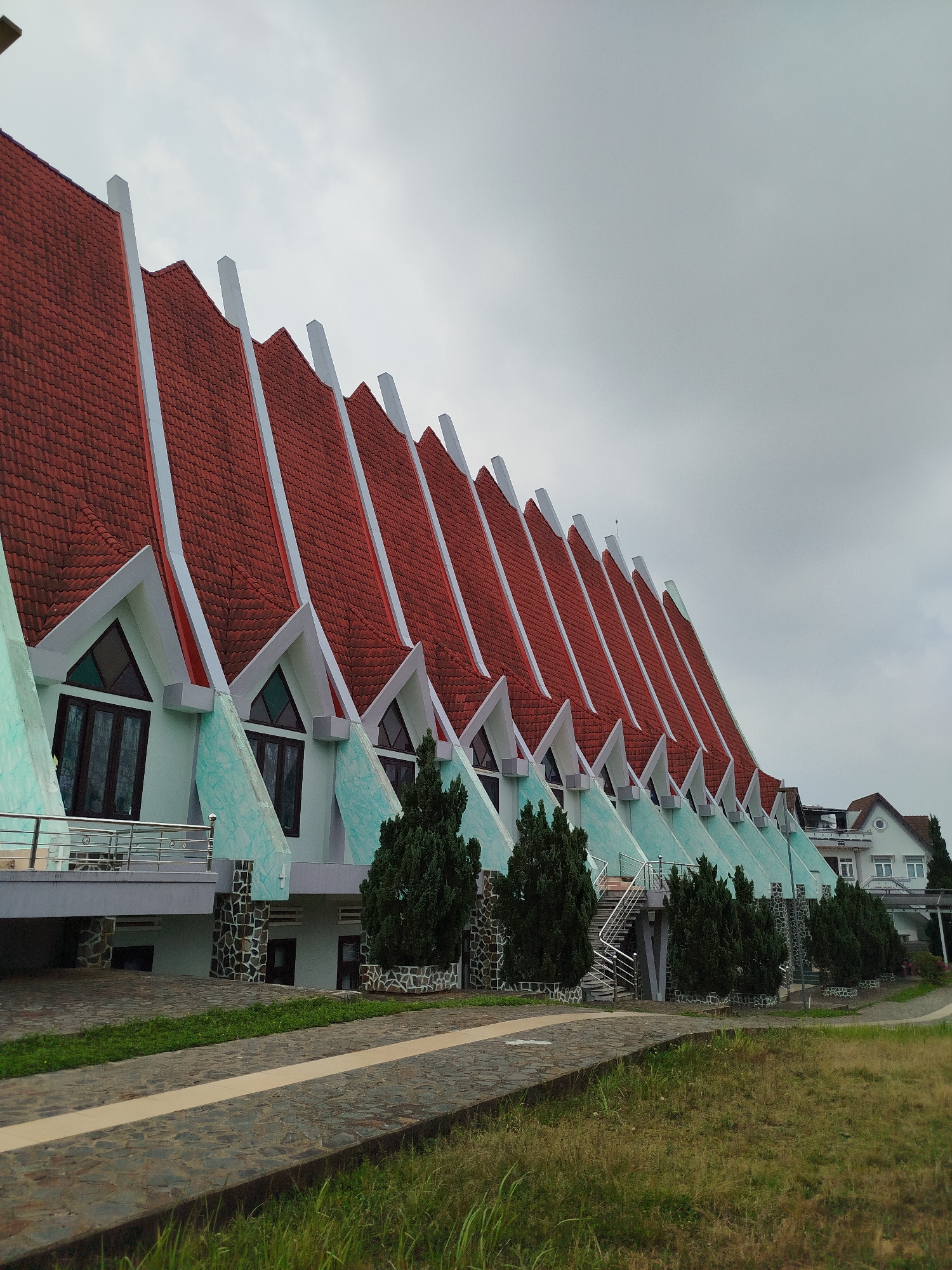 The image shows a building in Lâm Viên - Đà Lạt, Vietnam. The building has a distinctive, repeating gabled roofline with red tiling and white trim. The walls are a light green with a mottled texture. There are windows under each gable. Evergreen trees line the base of the building. A stone walkway is visible in the foreground, flanked by grassy areas. In the background is a white building. The sky is overcast.