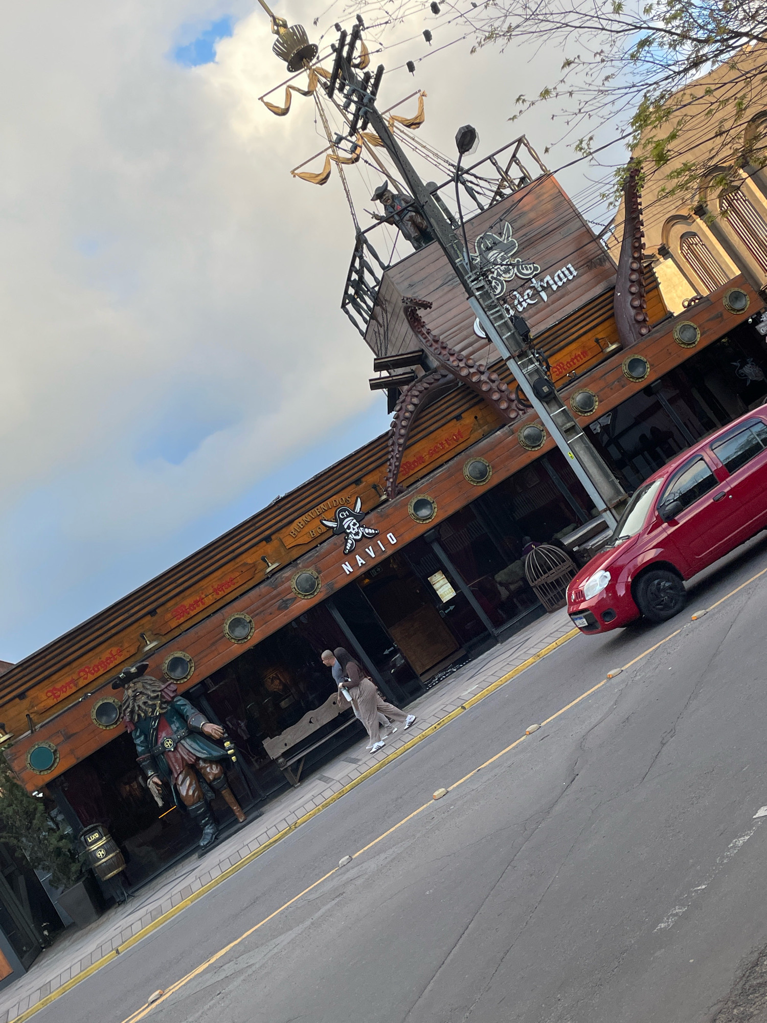 The image depicts a street scene in Gramado, Brazil, dominated by a building designed to resemble a pirate ship. The structure features wooden planks, porthole windows, and a mast with rigging and decorative sails. A large, detailed statue of a pirate stands prominently in front of the establishment. A red car is parked on the right side of the street, and two individuals are walking on the sidewalk in front of the pirate ship building. The sky above is cloudy with patches of blue. The building's signage indicates it is likely a restaurant or bar with a pirate theme.