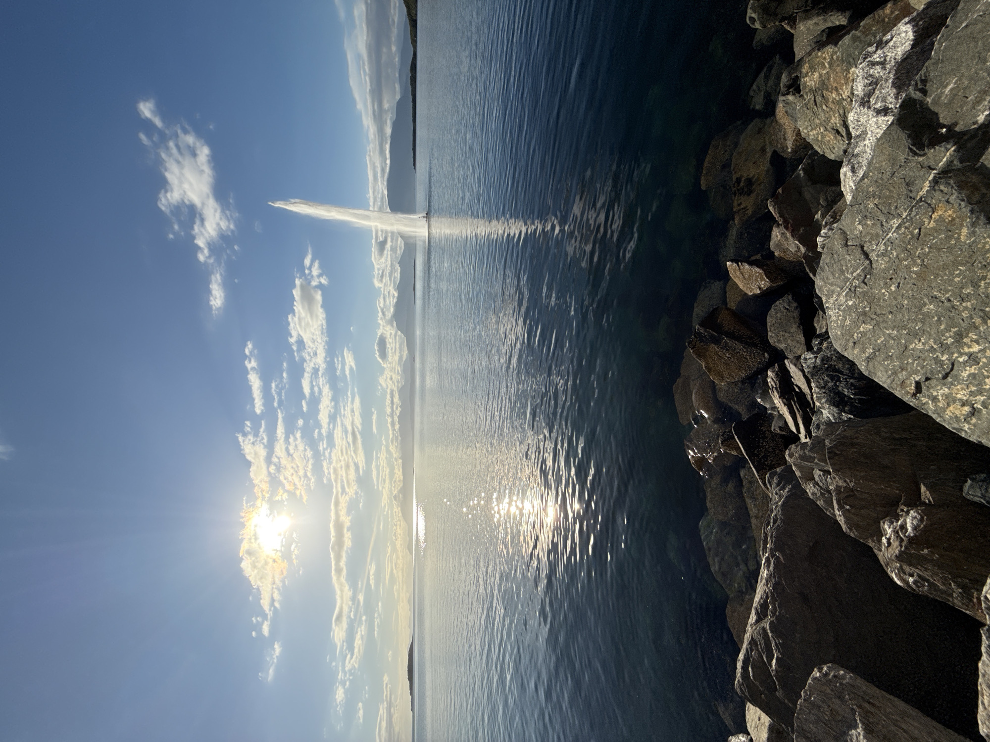 A large water fountain is erupting in the distance, shooting a tall plume of water into the sky. The fountain is located in the sea, with landmasses and mountains visible on the horizon. The sun is shining brightly, casting a glare on the water and creating reflections. The foreground of the image is lined with large rocks, forming a natural barrier along the water's edge. The sky is blue with scattered clouds. The scene appears to be set in Harstad, Norway, a coastal city known for its natural beauty.