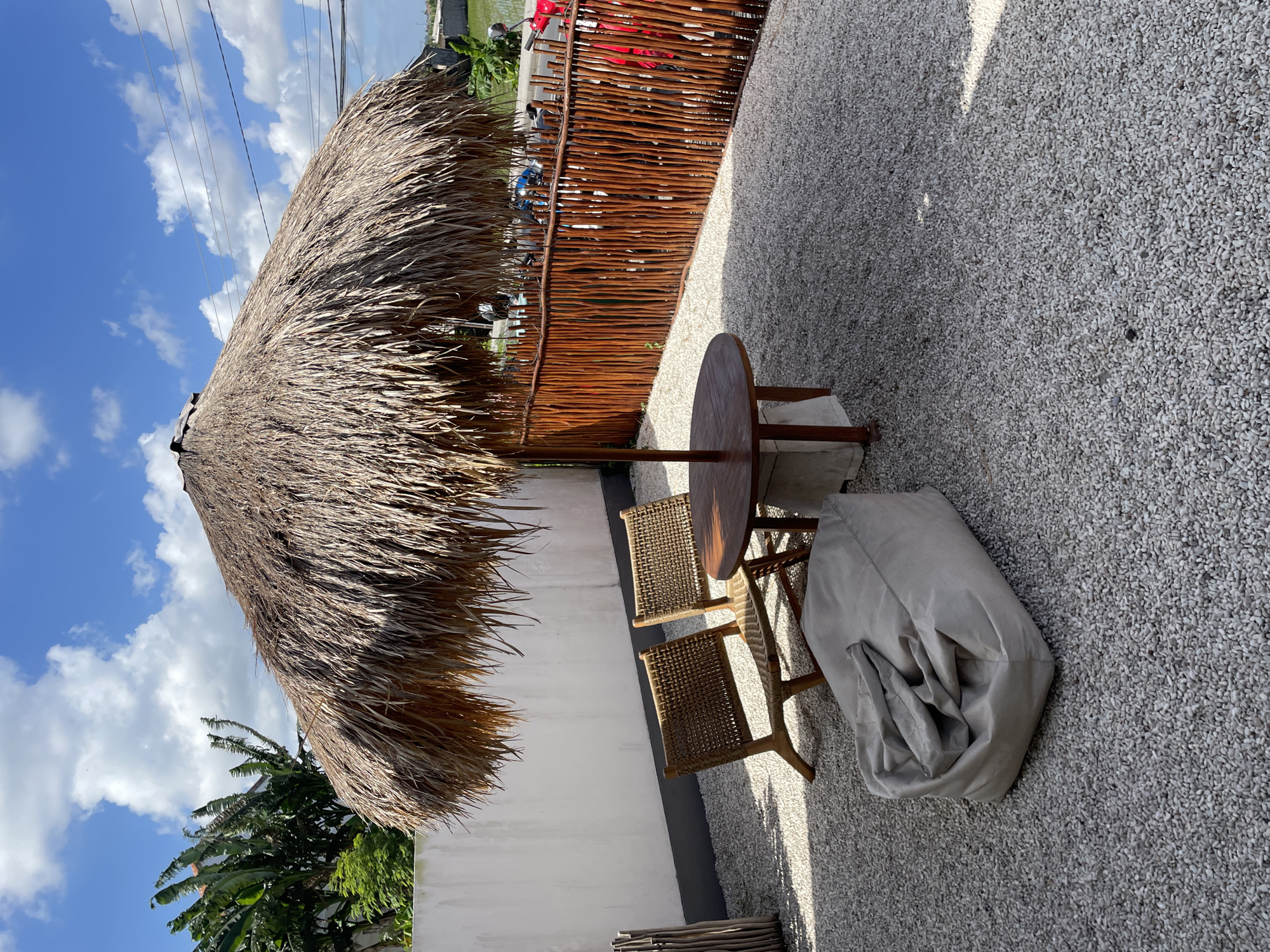 An outdoor seating area is depicted, located in Mengwi, Indonesia. The area is covered with fine white gravel. A large thatched umbrella provides shade for a circular wooden table and two woven chairs. In front of the table and chairs, a light-colored beanbag chair rests on the gravel. A rustic wooden fence made of thin vertical branches encloses the seating area. Beyond the fence, several motorcycles are visible, along with a glimpse of lush green vegetation and a blue sky with scattered white clouds. A white wall with a dark trim runs along the left side of the frame, and banana trees are visible behind it. The lighting suggests it is daytime, with shadows cast by the umbrella and furniture.
