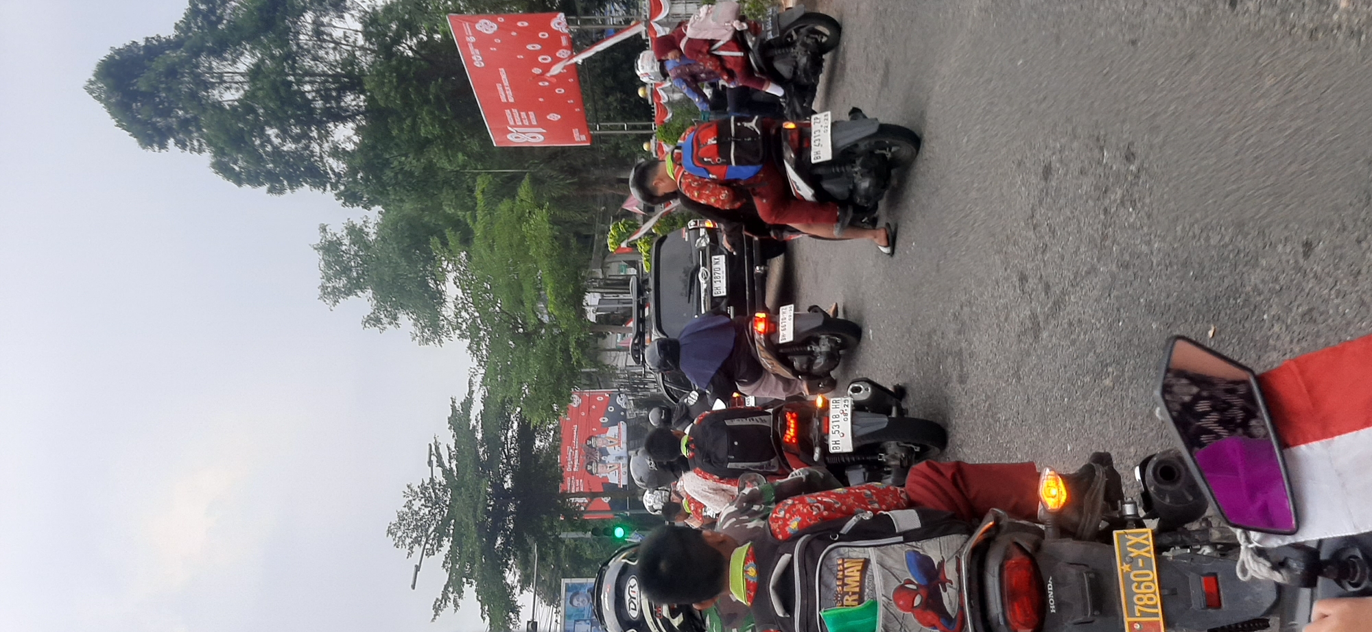 The image shows a traffic scene in the city of Jambi, Indonesia, on a cloudy day. Several motorcycles and a car are visible on a paved road. Many individuals are riding on the motorcycles, some of whom appear to be children wearing red and white school uniforms and backpacks. One motorcycle in the foreground has a license plate reading "7860-XX." In the background, there are trees and a large red billboard with white text and graphics. The billboard appears to be a celebratory advertisement, possibly for an anniversary, with the number "81" prominently displayed. Traffic lights and street signs are also present. The general atmosphere suggests a busy street with ongoing traffic, possibly during a commute or a public event.