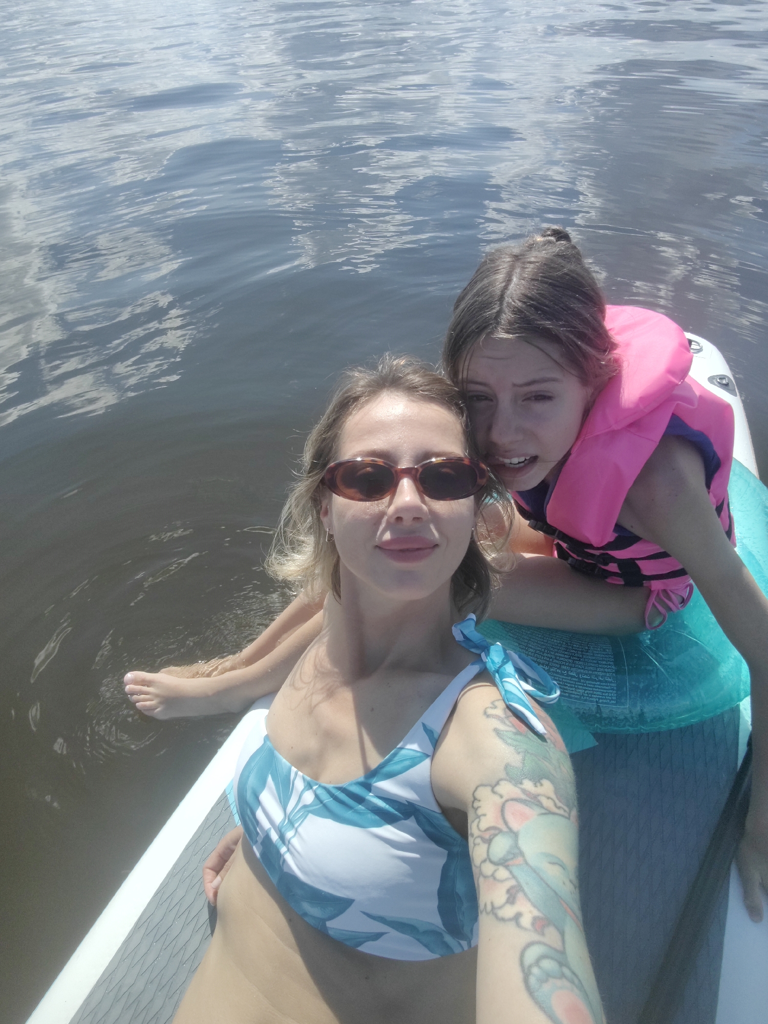 Two individuals are on a paddleboard in the water. The person in the foreground is a woman with blonde hair, wearing sunglasses and a bikini with a blue and white floral pattern. She has a tattoo on her arm. To her right and slightly behind her is a younger person, likely a child or teenager, wearing a pink life vest. They are both looking towards the camera. The water appears to be a lake or calm body of water, reflecting the sky. The scene suggests a recreational activity on a sunny day. The location is not identifiable as Saint Petersburg, Russia based on the visual information in the image.