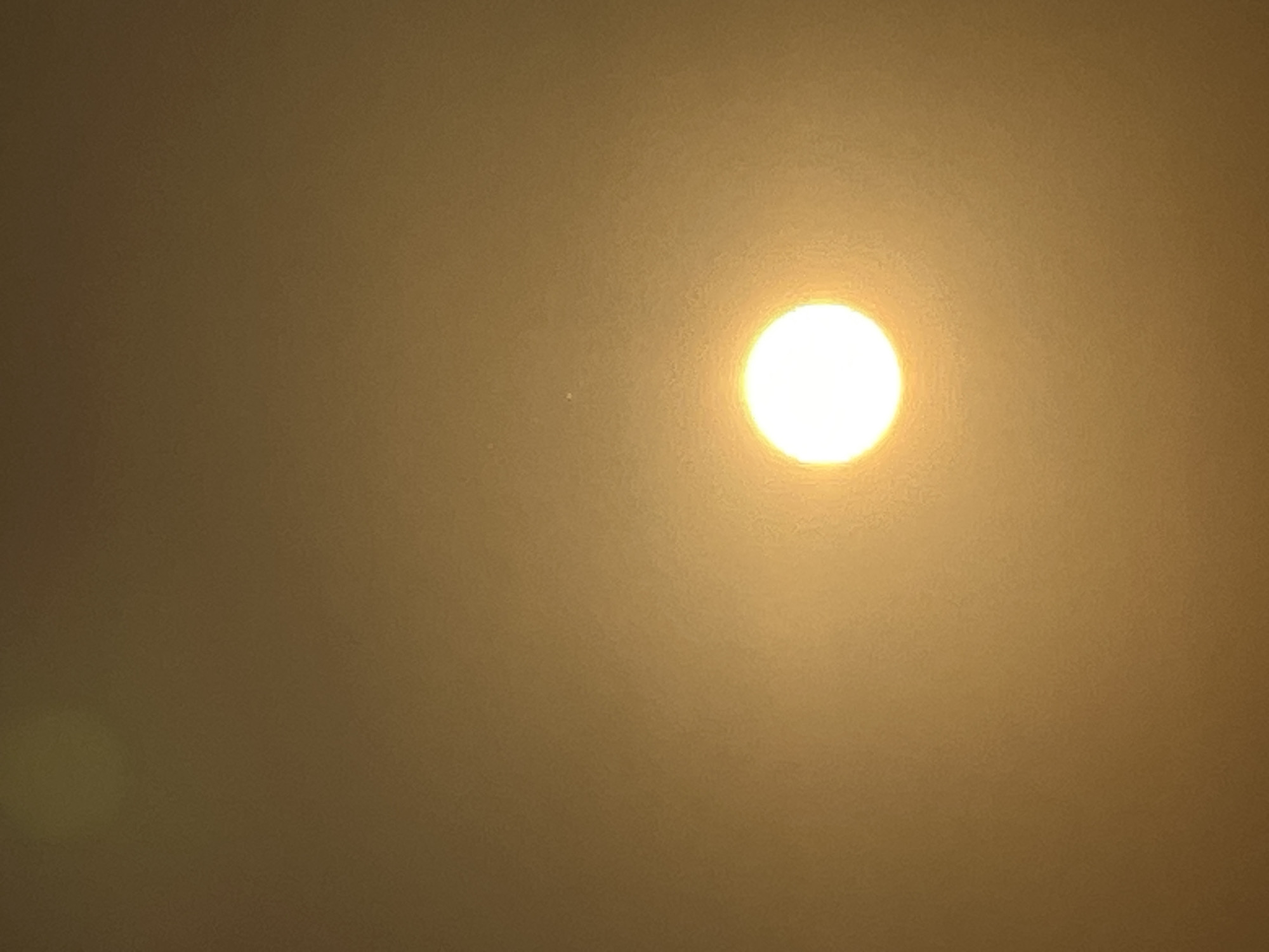 The image shows a bright, circular light source, likely the sun, dominating the center of a muted, yellowish-brown background. The background appears hazy or dusty, with a slight gradient of brightness, being lighter around the central light source. There is a faint, out-of-focus circular blur in the upper left corner of the image, possibly another light source or a lens flare. The overall impression is that of a very bright object seen through atmospheric conditions, such as smog, haze, or dust, common in urban or industrial environments. There are no identifiable objects, people, or interactions visible in the scene.