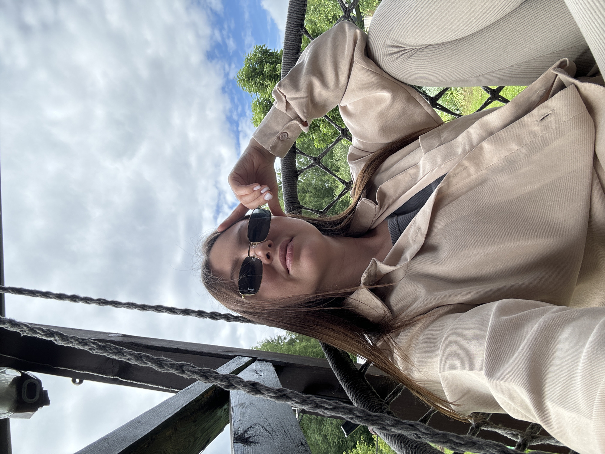A young woman with long brown hair, wearing sunglasses and a beige button-up shirt over a grey top, is seated in a hanging chair outdoors. She is looking at the camera with her hand resting on her forehead. The chair is suspended by thick ropes and appears to be on a porch or balcony with wooden beams above. The background shows green trees and a cloudy sky. The location is unspecified but likely in a rural or suburban setting, given the natural surroundings.